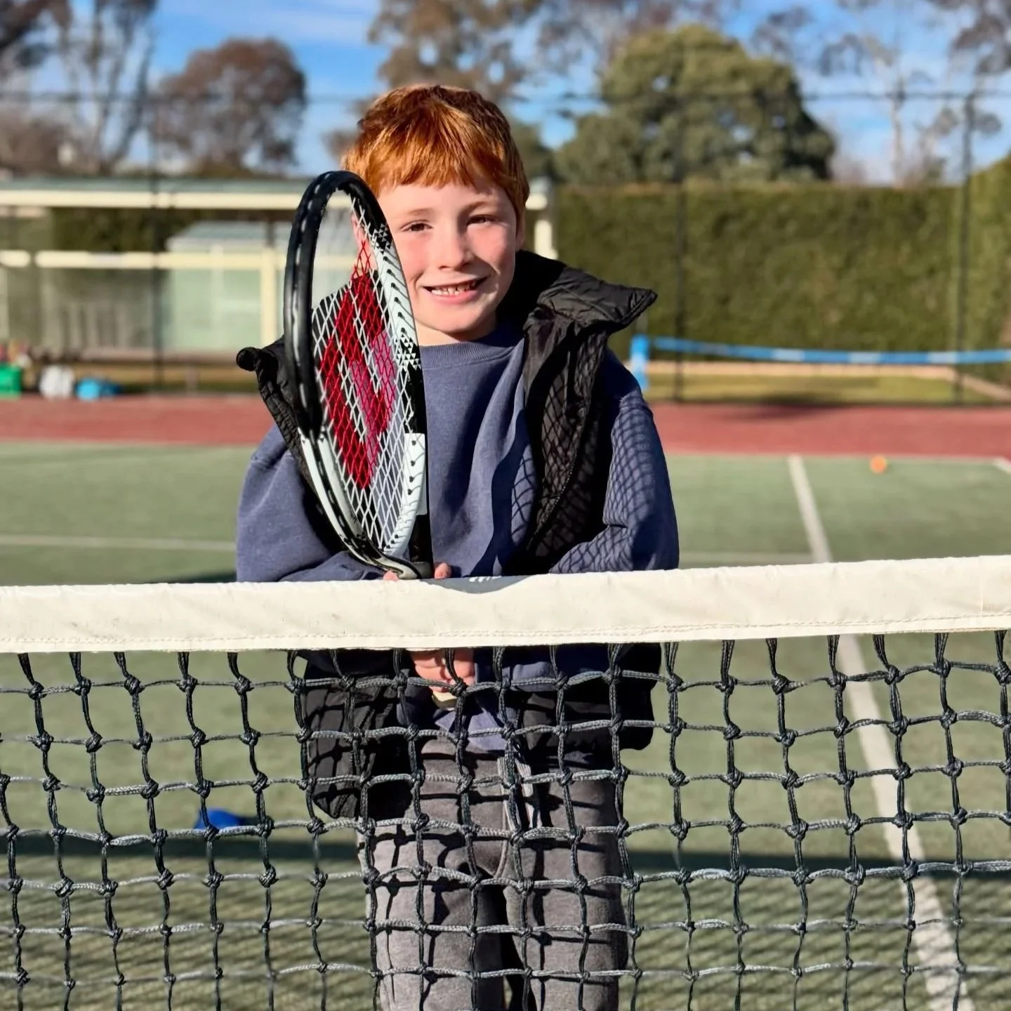 A young boy with red hair smiles on a tennis court, holding a tennis racket, standing behind the net.