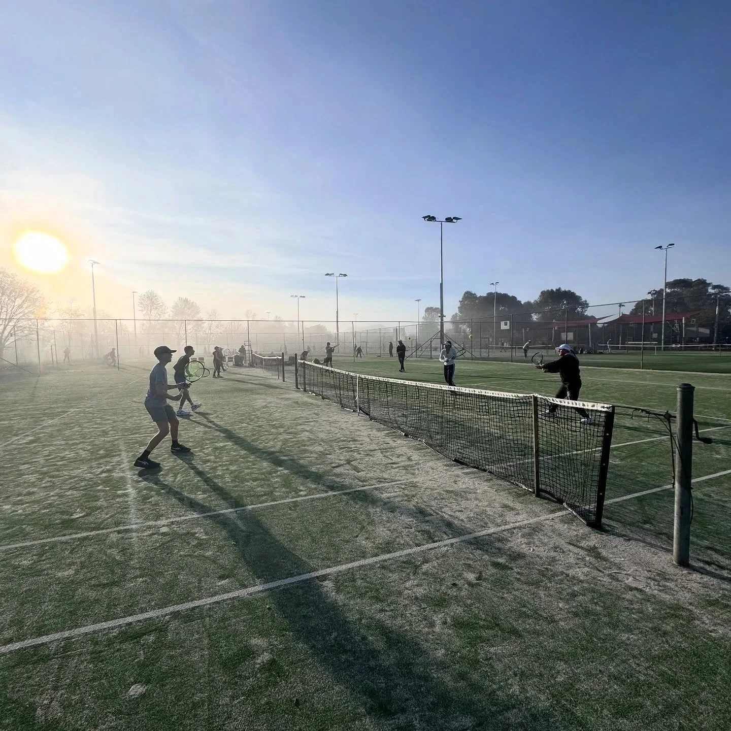 Tennis players play on outdoor courts with a cloudy sky and sunlight, surrounded by trees and tall lighting poles.
