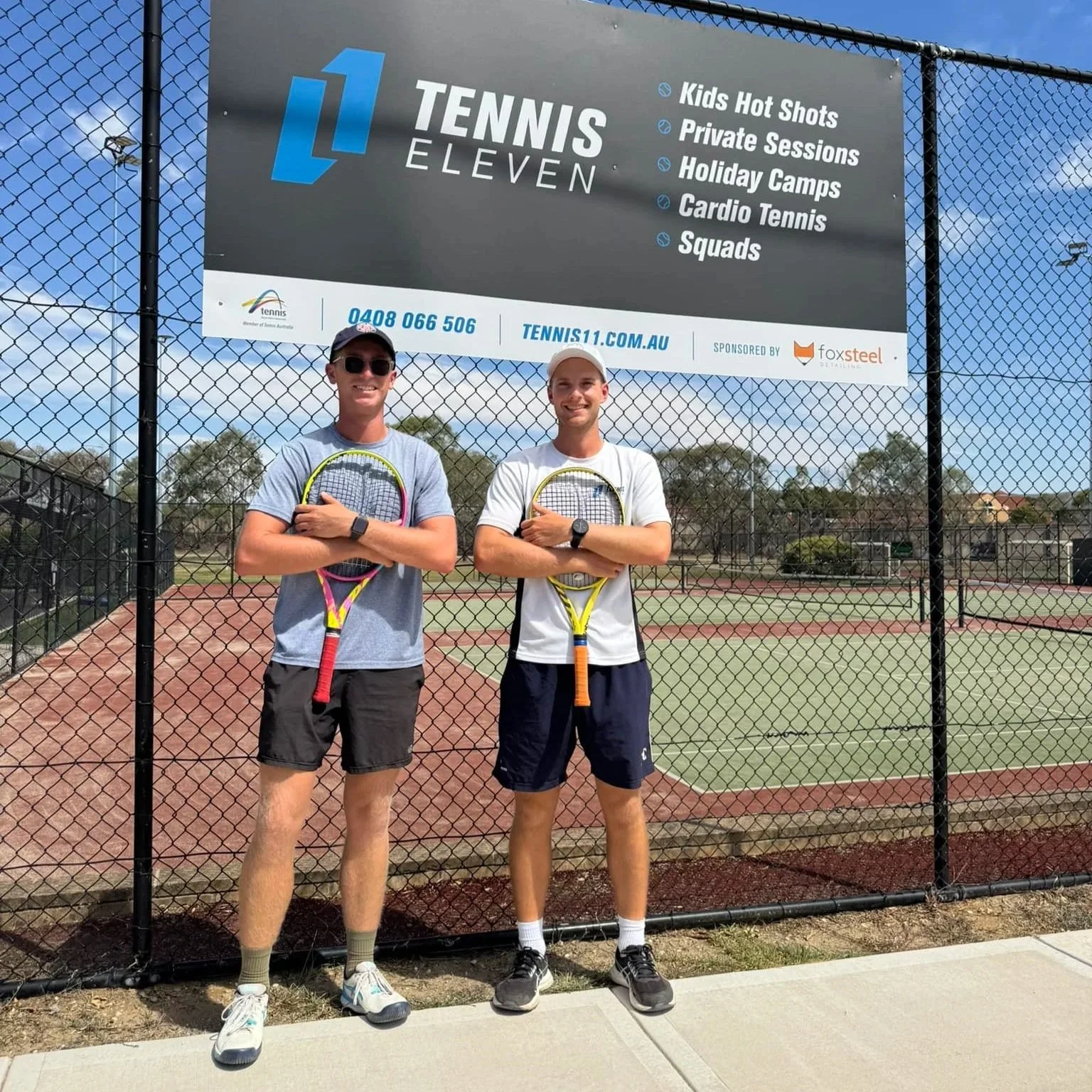 Two young men with crossed arms holding tennis racquets at a tennis court, standing in front of a large sign advertising Tennis Eleven with listed services, under a partly cloudy sky.