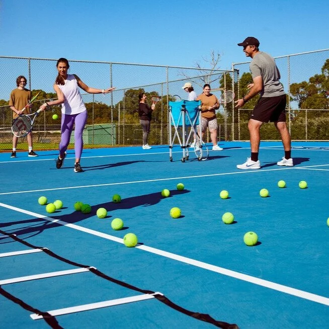 Adult players taking part in a group tennis coaching class with drills and ball exercises on an outdoor court.