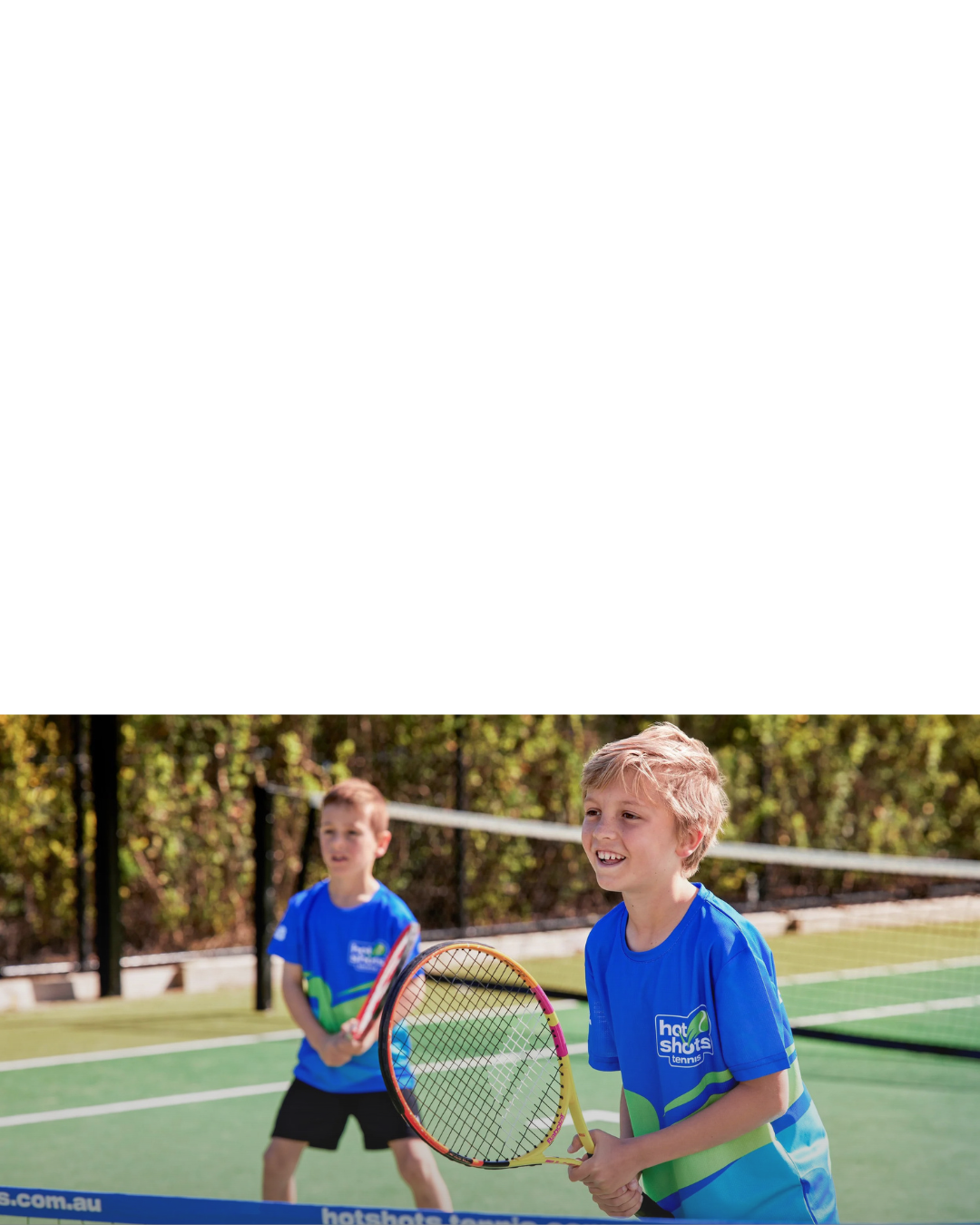 Two young boys wearing blue Hot Shots uniforms stand at a tennis net with racquets in hand.
