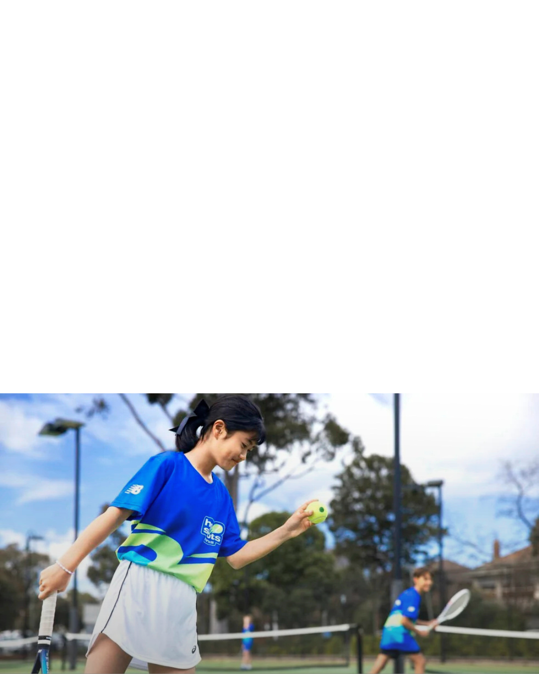 A young girl in a blue Hot Shorts shirt and white tennis skirt holding a tennis ball in her left hand and a tennis racquet in her left hand on a tennis court.