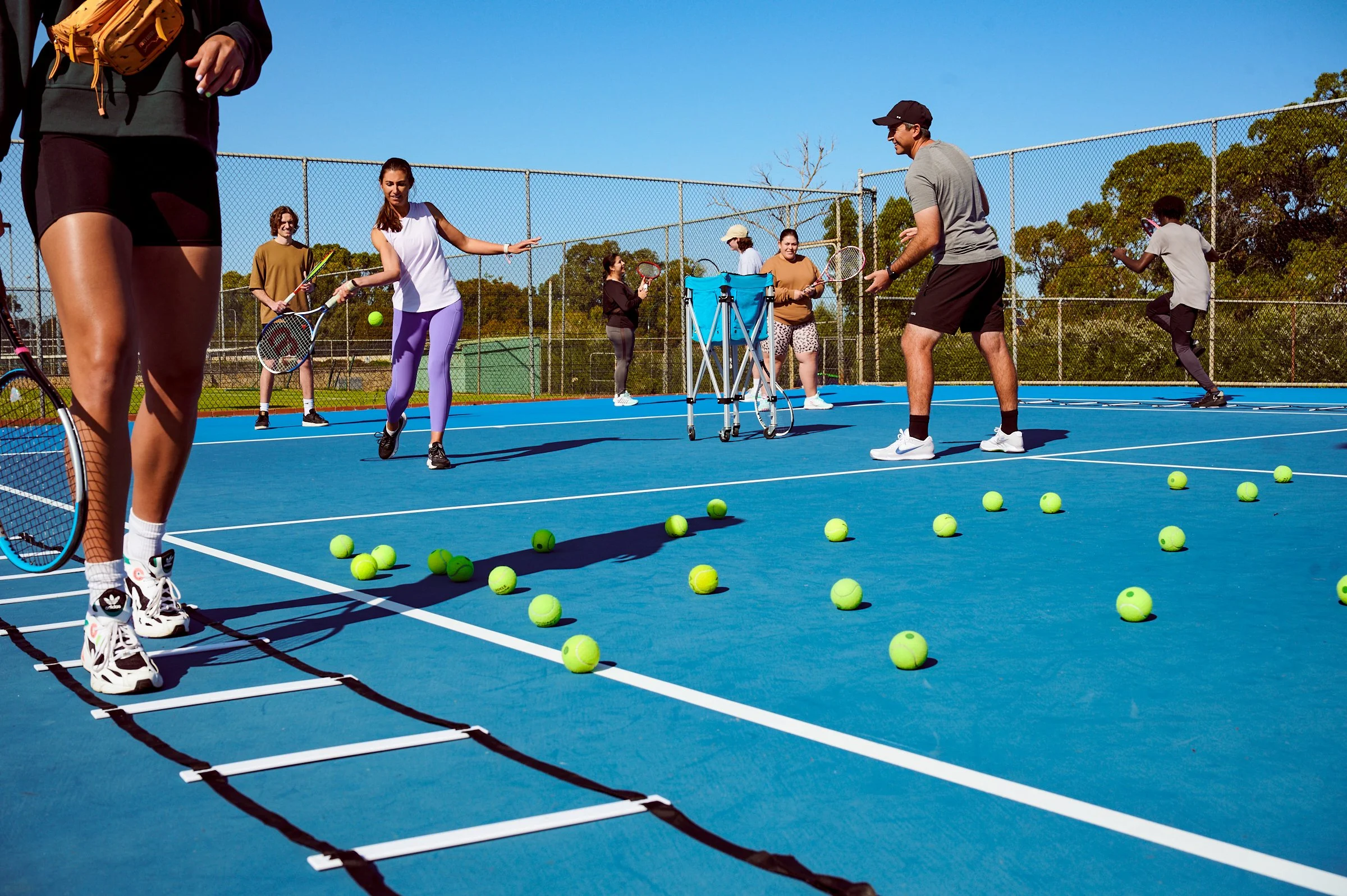 Adult players taking part in a group tennis coaching class with drills and ball exercises on an outdoor court.