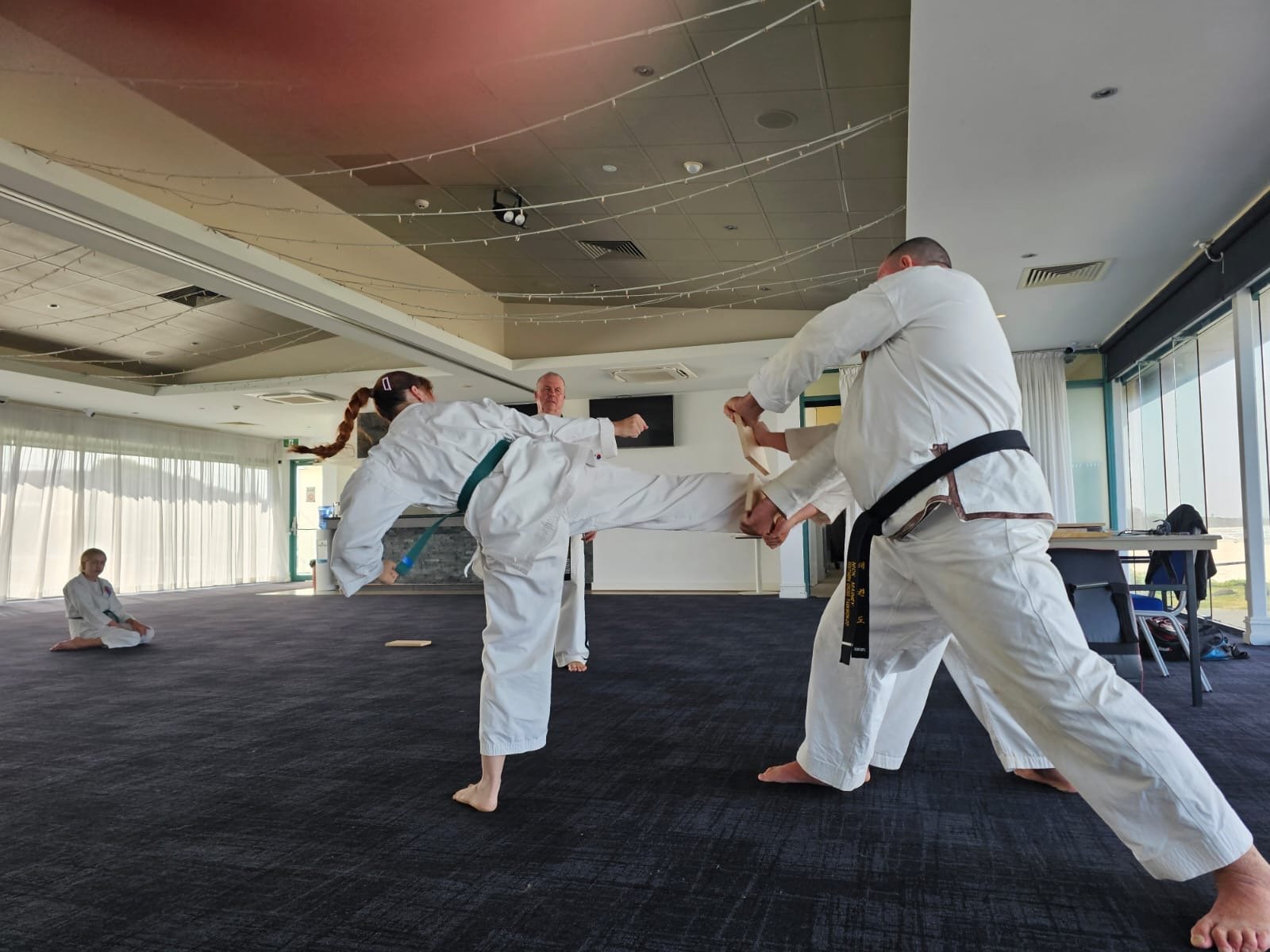 Two martial artists practicing a kick in a dojo, with a child watching in the background.