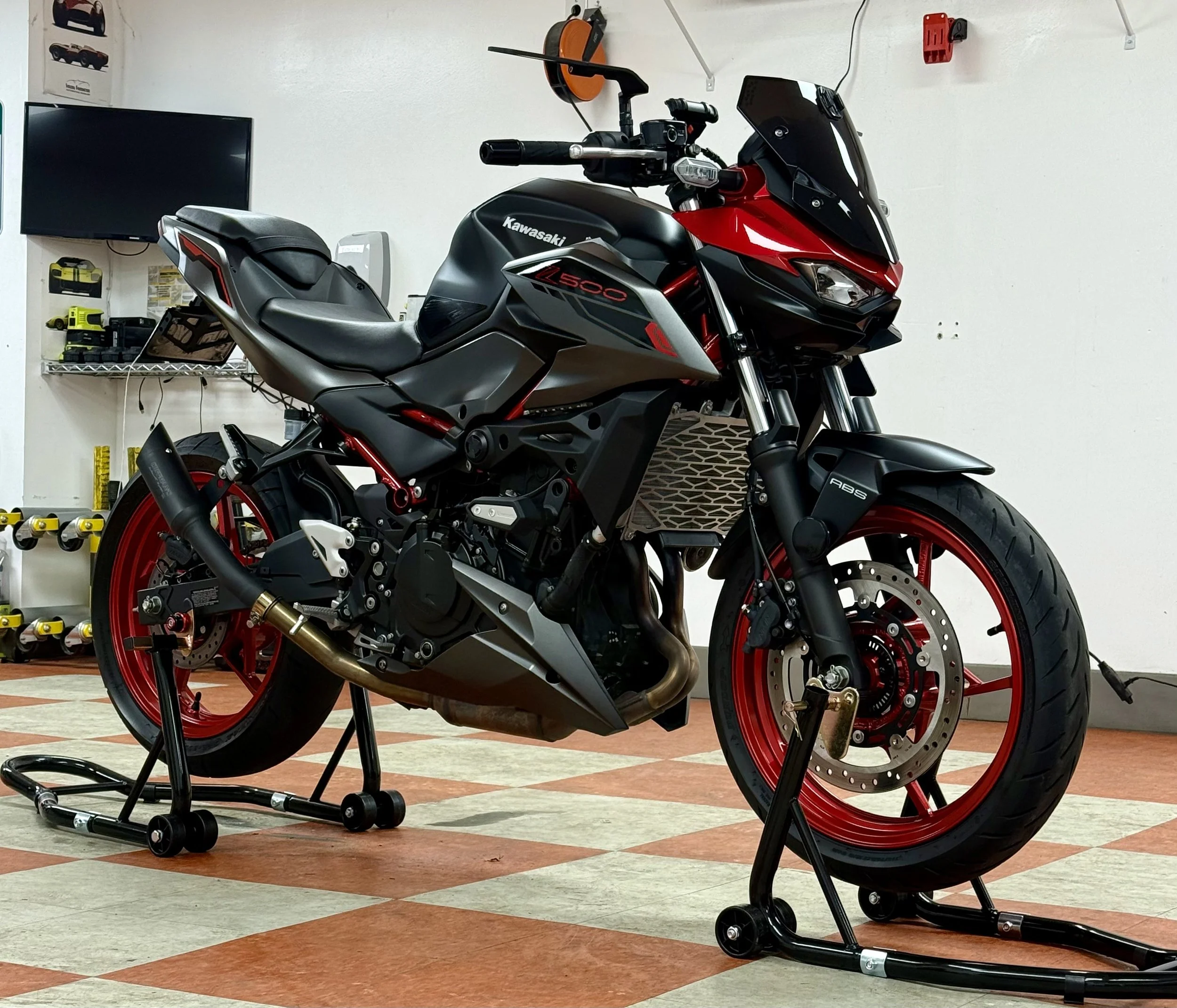 A Kawasaki sport motorcycle is set on a stand indoors, featuring a black and red color scheme with red wheels, a black exhaust, and a black windshield. The background includes a wall with tools, a TV, and shelves.