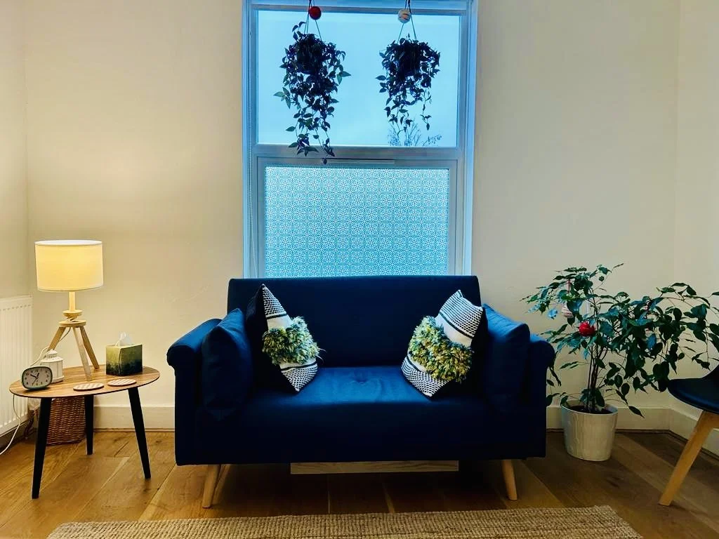 A cozy living room corner with a navy blue sofa, decorative pillows, a wooden side table with a lamp, a small clock, and a tissue box. There is a potted plant and a black chair next to it, with a window behind the sofa, decorated with hanging plants.