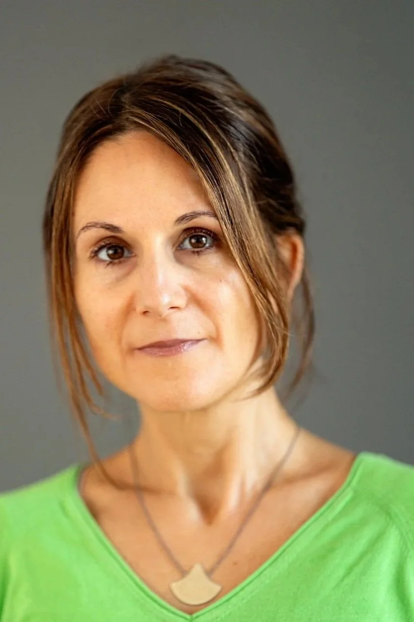Close-up portrait of a woman with short brown hair and a green shirt, looking at the camera with a neutral expression, against a plain gray background.