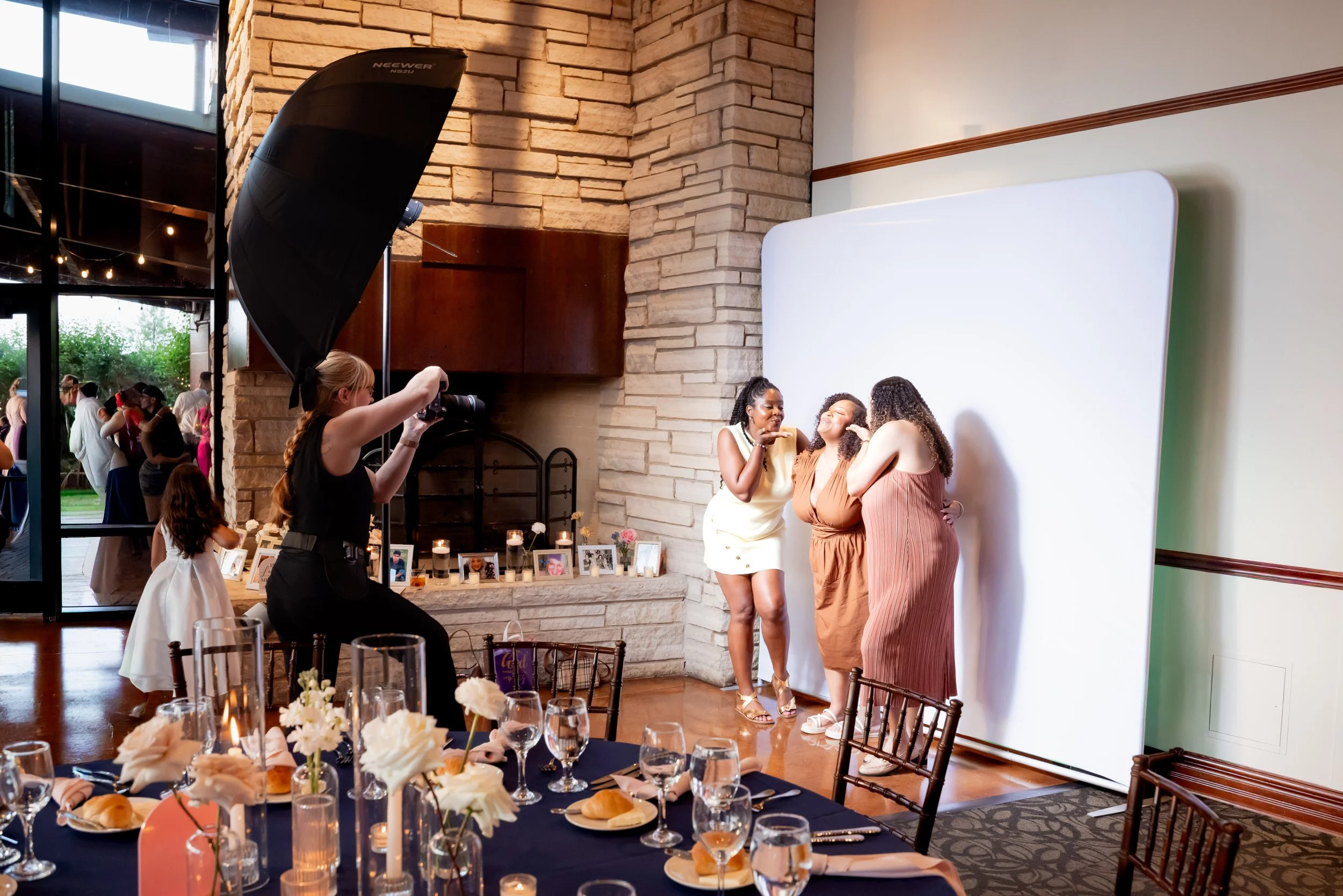 Three women posing for a photo in front of a white backdrop at an indoor event, with a photographer taking their picture. The setting includes a table with a black tablecloth, floral centerpieces, glasses, and tableware, and a fireplace with framed photos and candles in the background.