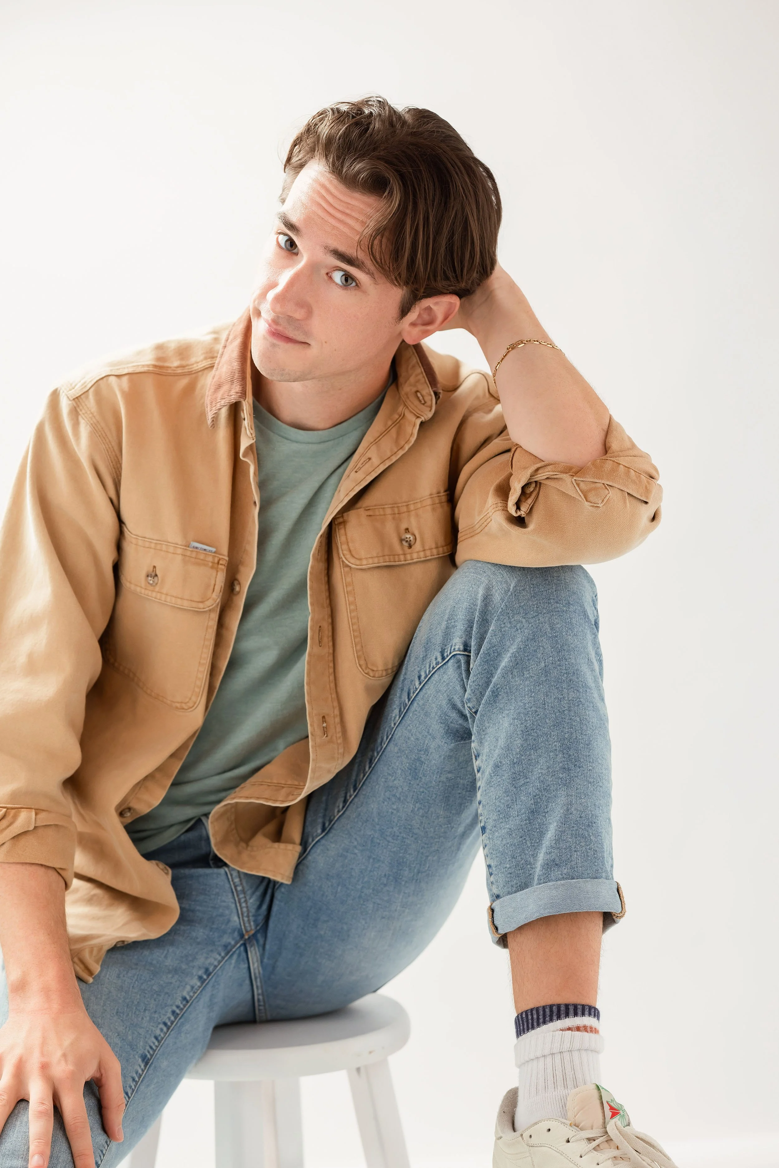 A young man sitting on a white stool with one leg crossed over the other, wearing a tan jacket, a light green T-shirt, light blue rolled-up jeans, a white sneaker, and a striped sock. He has brown hair and is looking at the camera with a slight smile, resting his head on his hand.