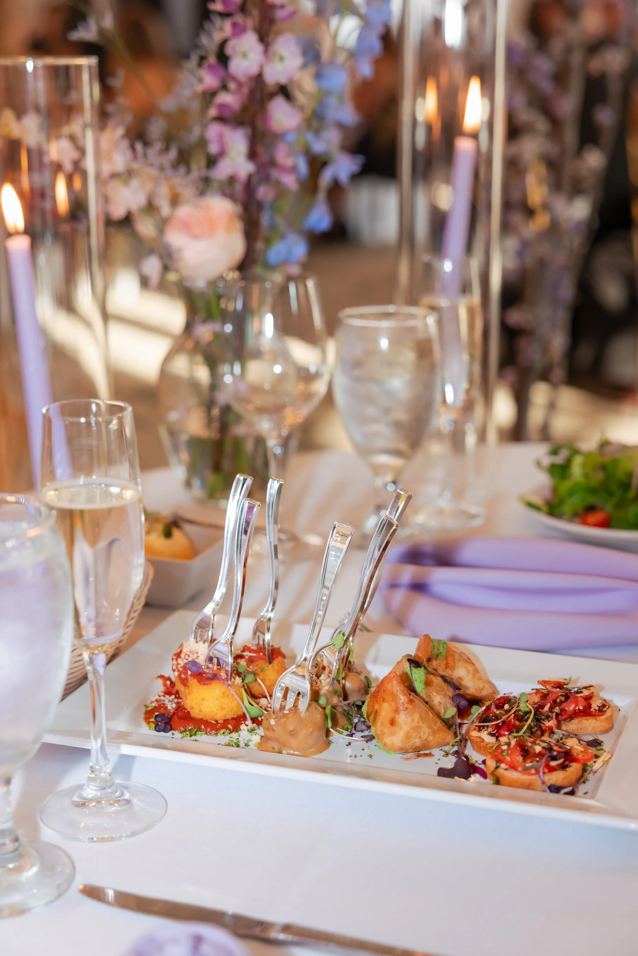 Plated appetizers on a table with candles, glassware, and floral centerpieces in the background.