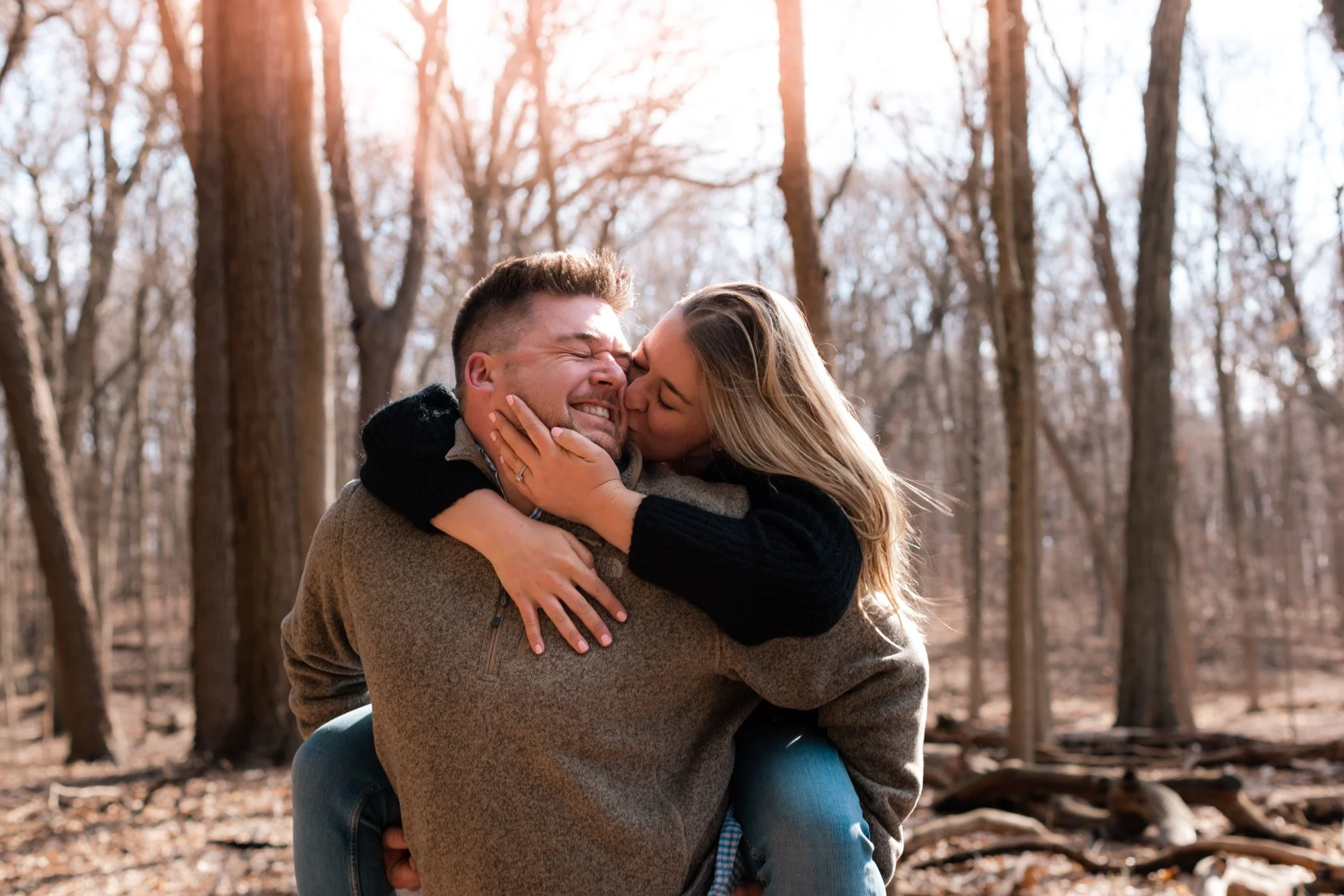 A couple happily embracing in a forest during the daytime, with sunlight shining through the trees.