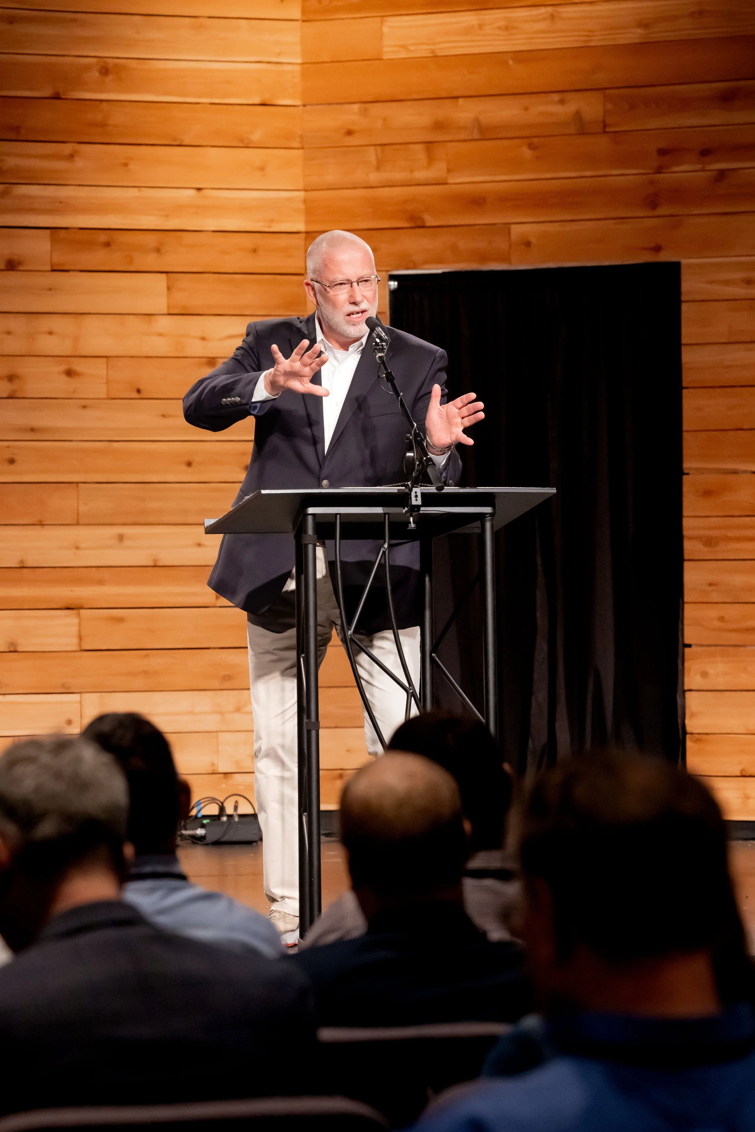 Speaker at a podium addressing an audience in a venue with a wood-paneled wall