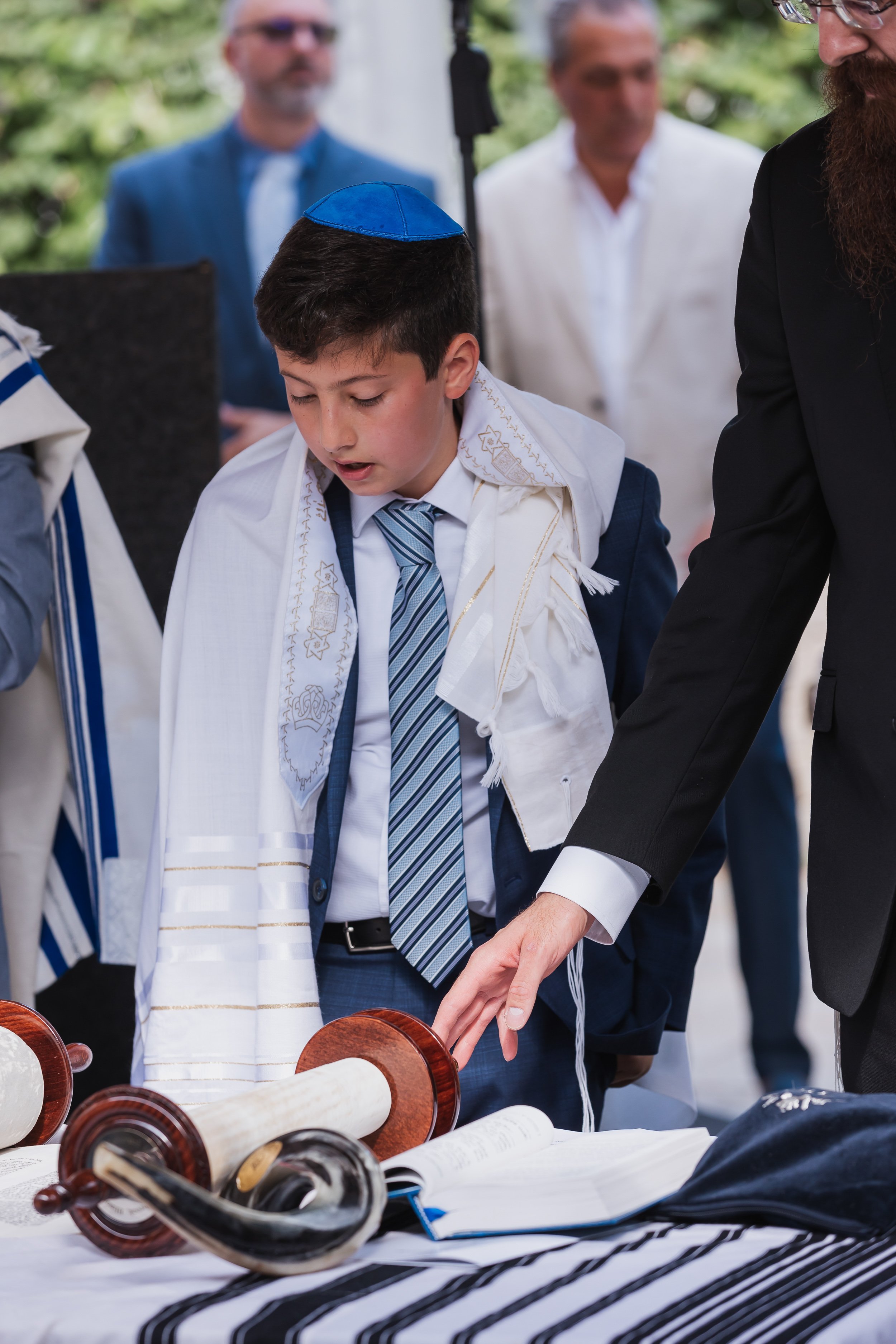 Boy wearing a tallit and kippah stands at a table during a bar mitzvah ceremony.