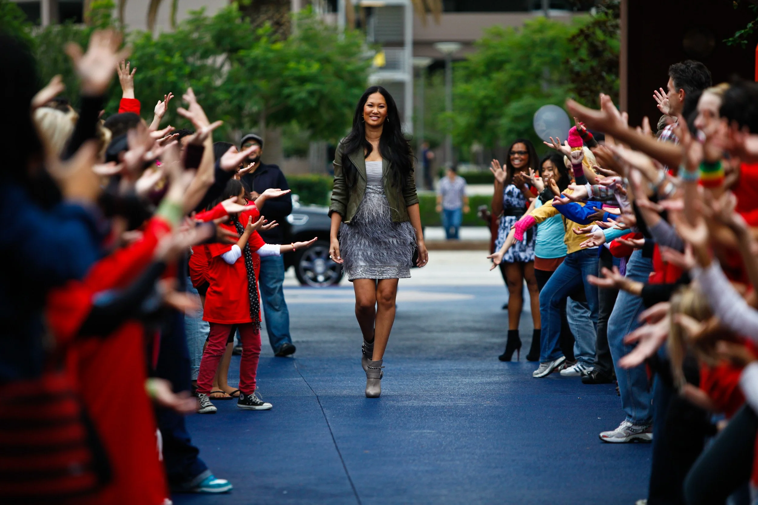A woman walking in the middle of a street, surrounded by people with outstretched arms, some taking photos, and children and adults on both sides, in an urban outdoor setting with trees and buildings in the background.