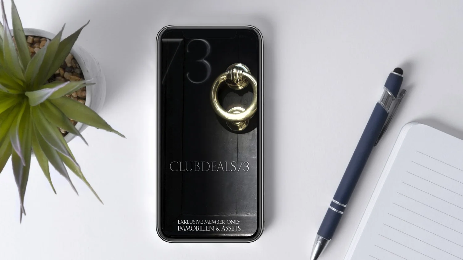 Smartphone mit goldener Türklopfer-Attrappe, daneben eine grüne Sukkulentenpflanze, ein blauer Kugelschreiber und ein weißes Notizbuch auf einem weißen Tisch.