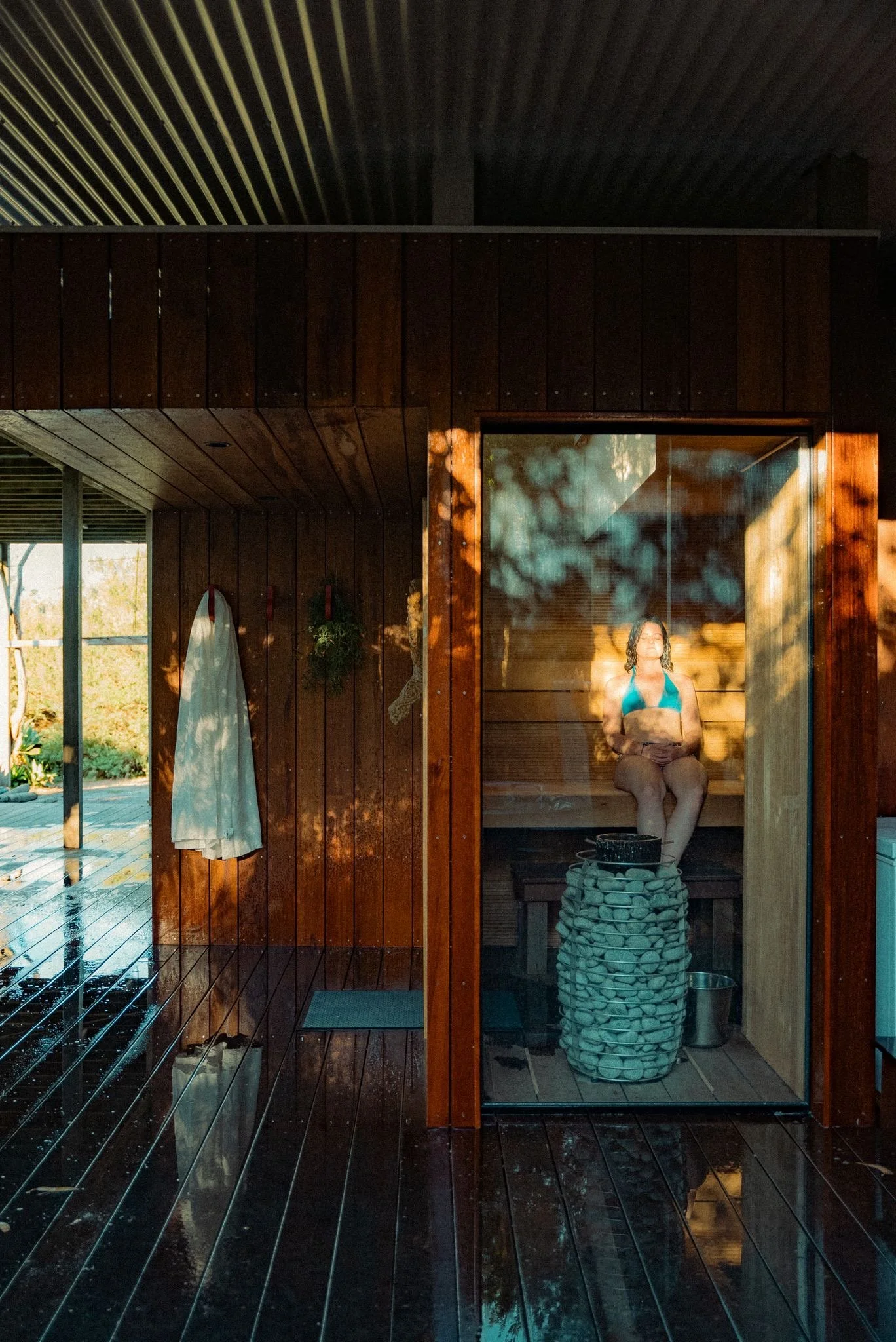A woman in a blue bikini sits inside a wooden sauna room, with sunlight creating shadows of nearby trees on the glass door and wooden wall.