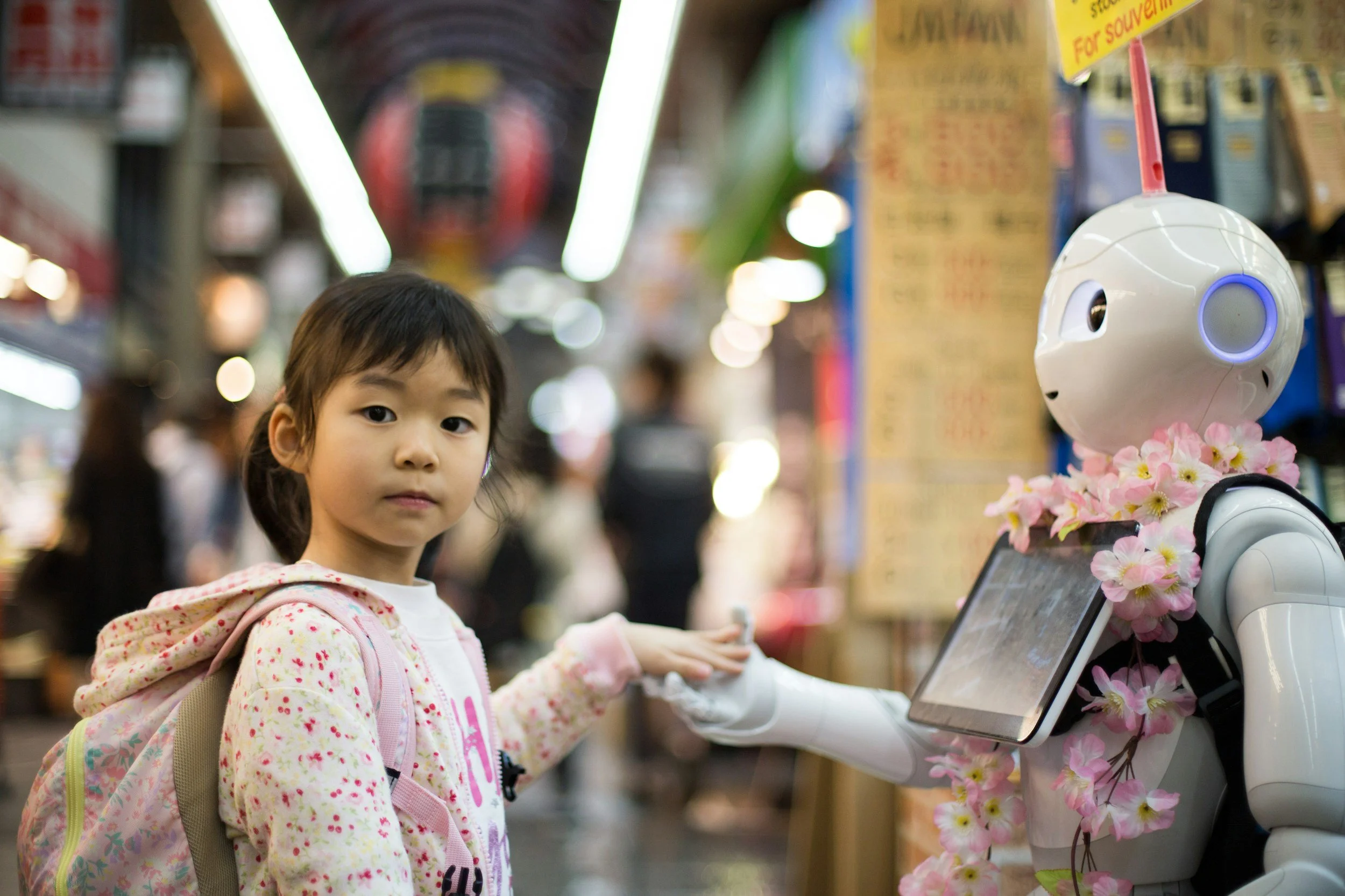 A young girl with a pink backpack touching a robot that has a tablet attached to its chest, in a busy indoor market or shopping area.