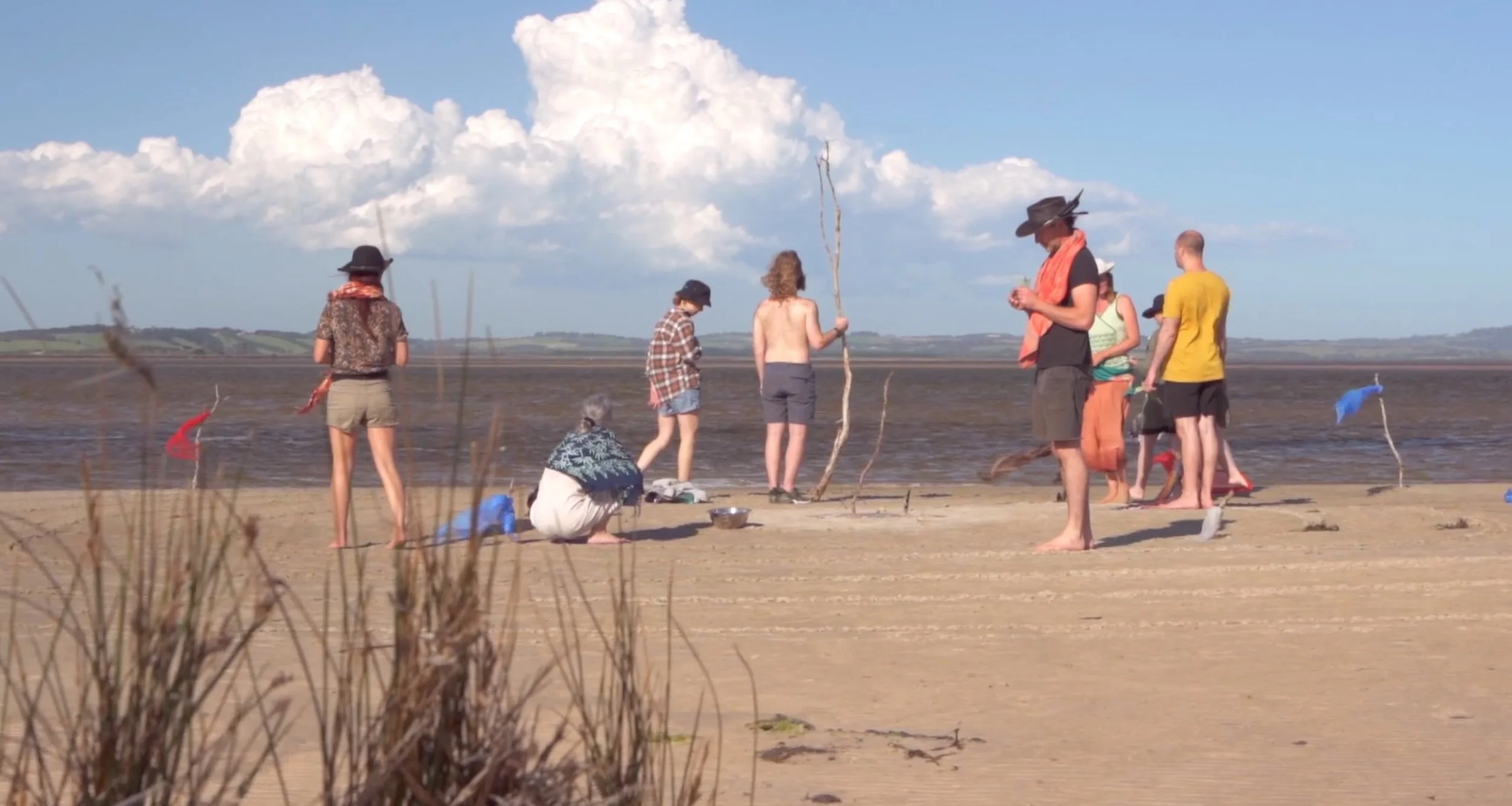 Group of people, including children and adults, gathered on a sandy beach with a body of water in the background, some dressed casually and some wearing hats, appear to be participating in a beach activity or gathering on a sunny day.