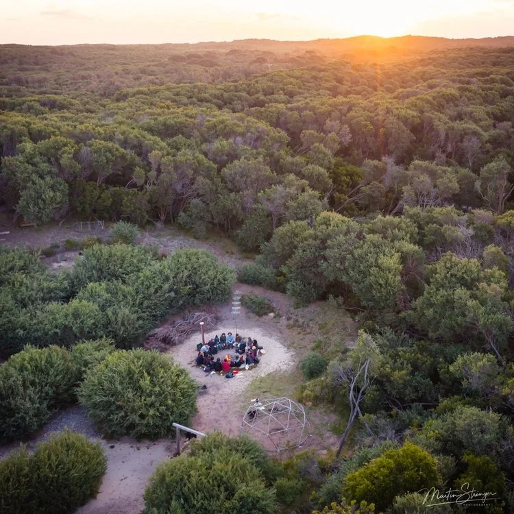 A group of people sitting in a circle around a campfire in a forested area during sunset.