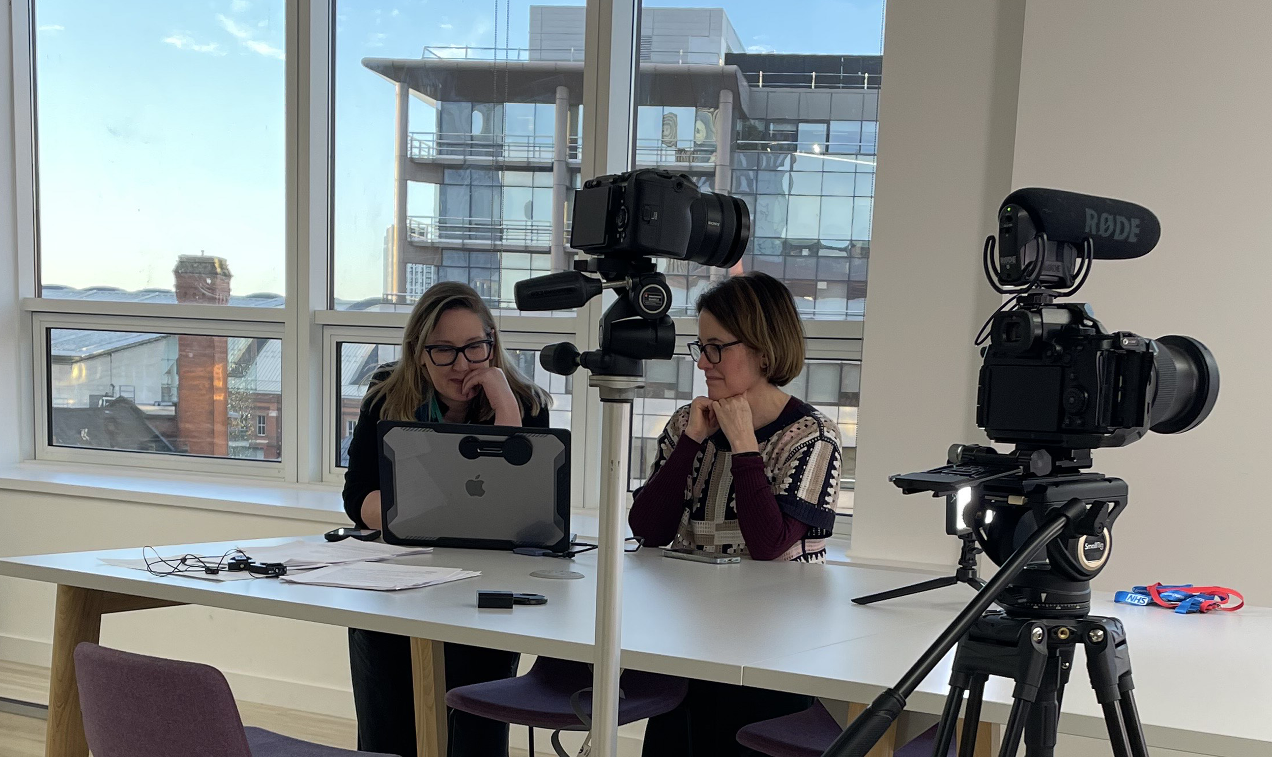 Two women sitting at a table with recording equipment, looking at a laptop, in front of large windows with city buildings in the background.