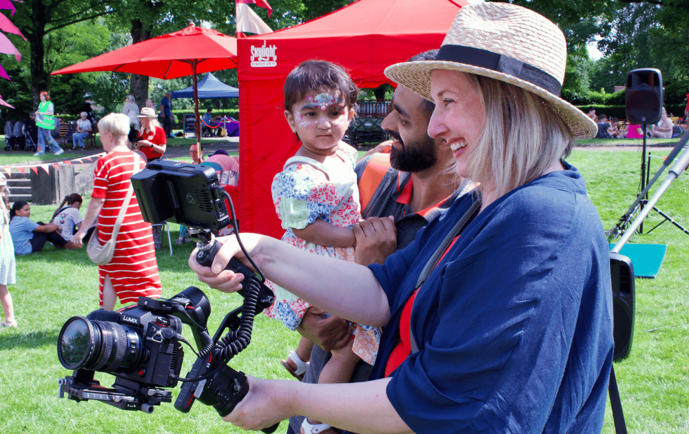 A woman, Kirstie Henderson, founder of Brave Day, wearing a straw hat and a blue jacket, films with a large camera in front of a man and a young girl at an outdoor event in a park. People and a red canopy are visible in the background.