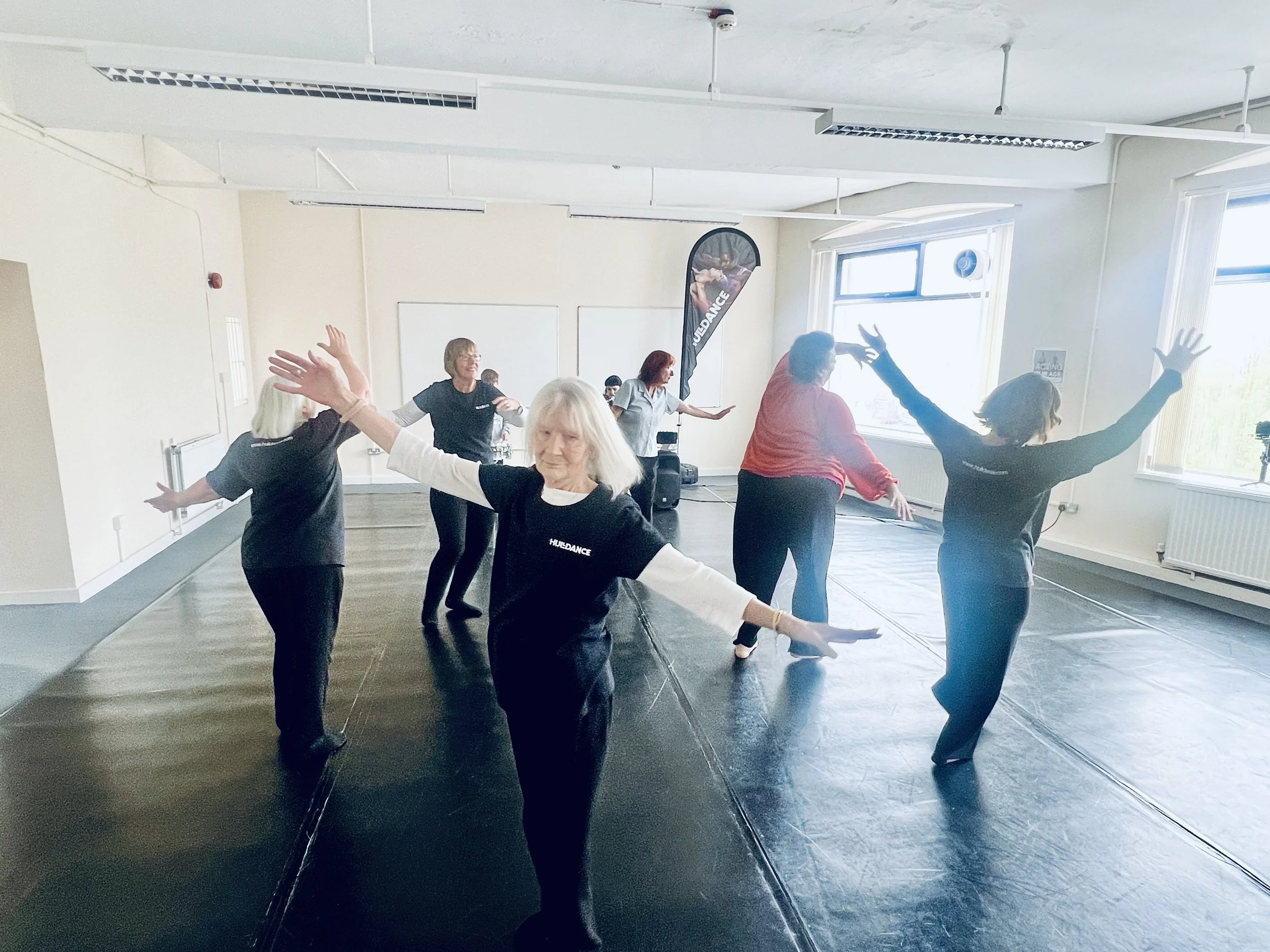 Group of women participating in a dance or exercise class in a bright studio with large windows, black mats, and a flag that reads "Husband".