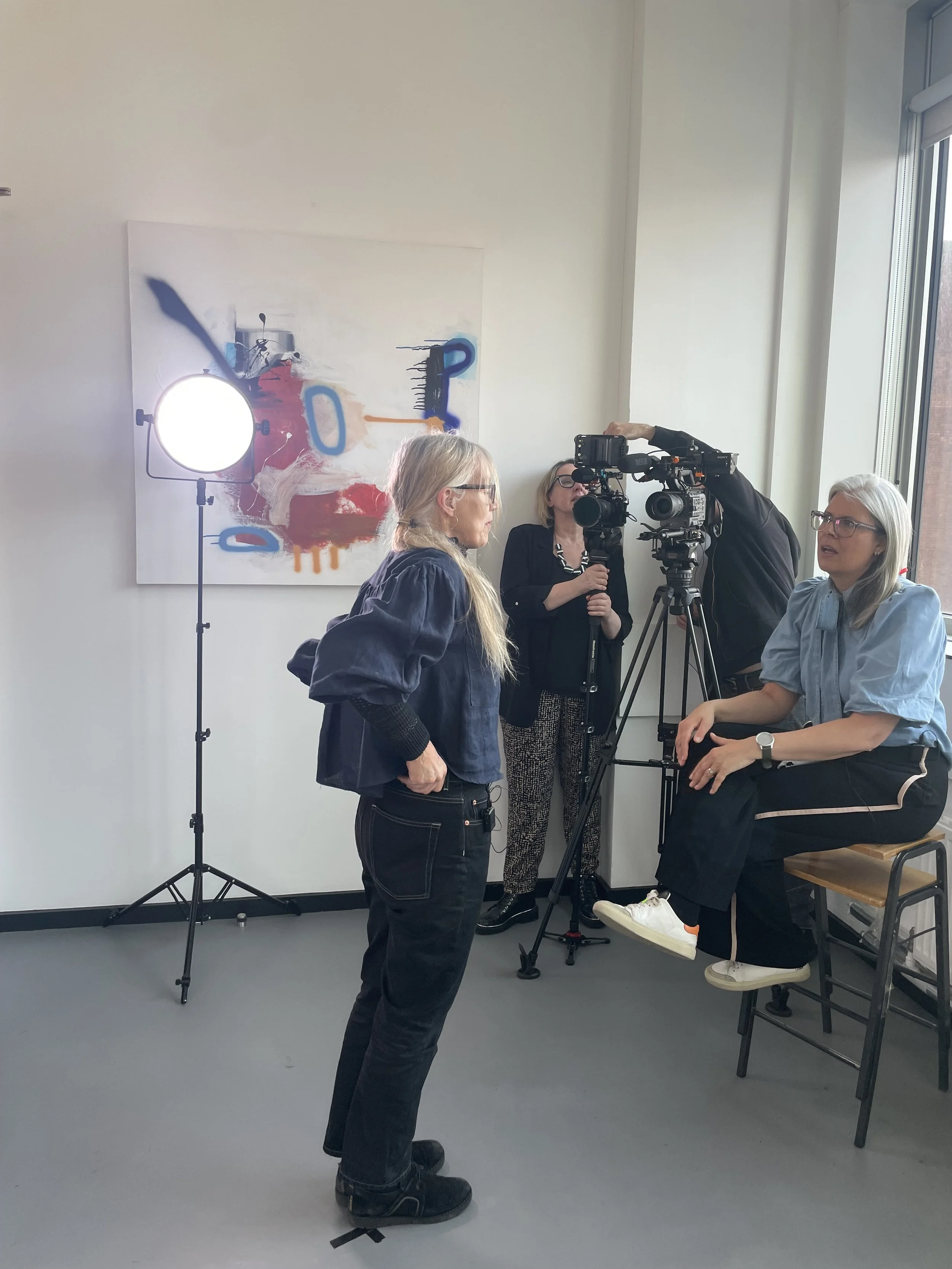A woman with long blond hair and glasses being filmed by a camera crew in a studio with a large colorful abstract painting on the wall and a bright light on a stand.