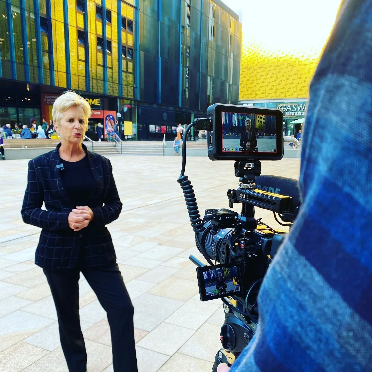 A woman being filmed outdoors in front of a modern glass building with golden panels, with a camera operator capturing her interview or speech.