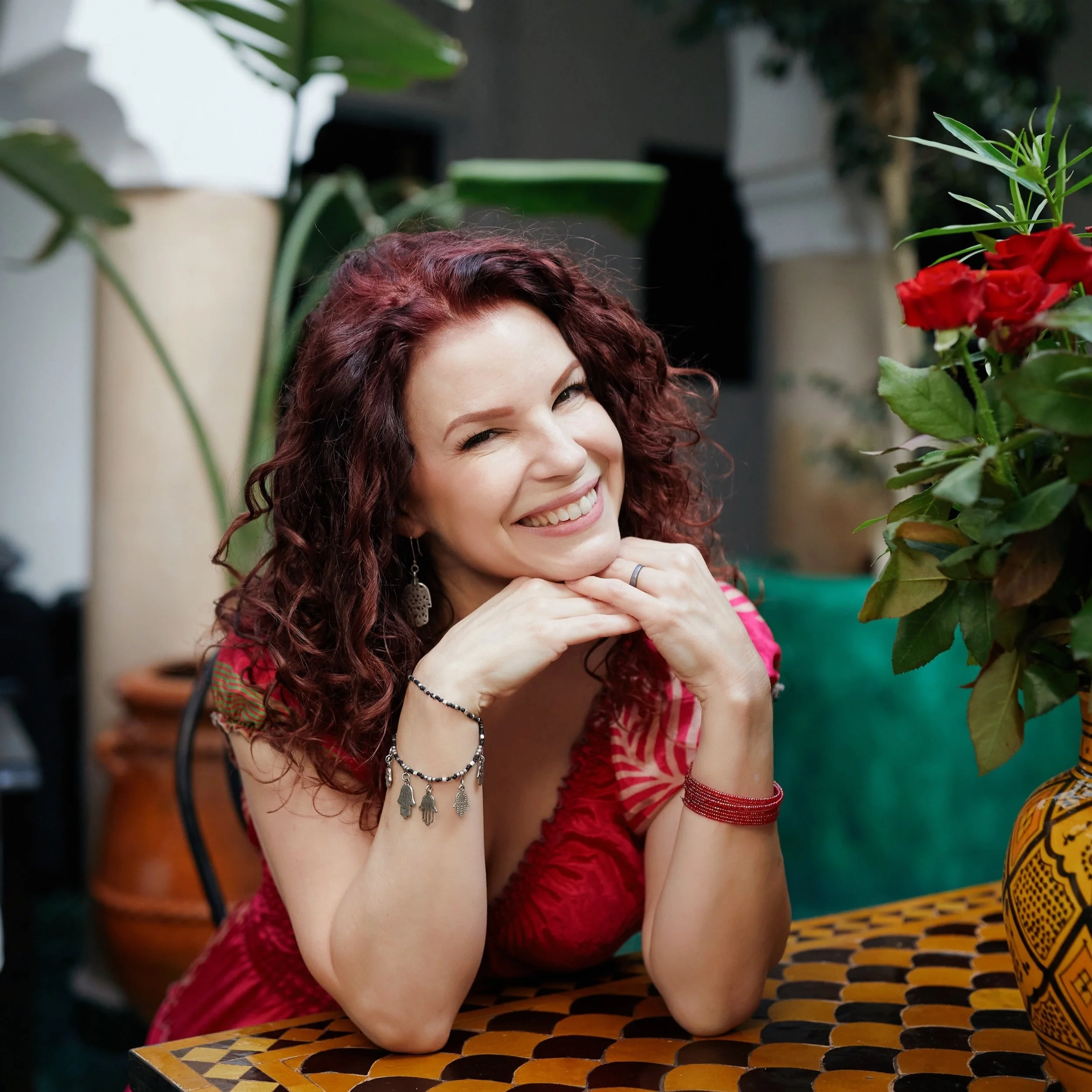 A woman with curly red hair smiling and posing with her chin resting on her hands at a table, surrounded by plants and flowers.