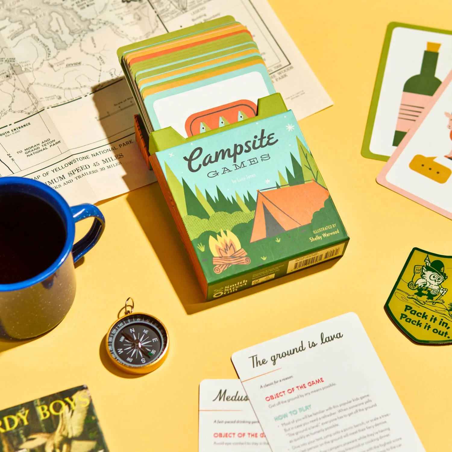 A camping-themed card game box with a tent and campfire illustration, a blue mug filled with a dark beverage, a compass, a handwritten card, and various game and map items on a yellow surface.