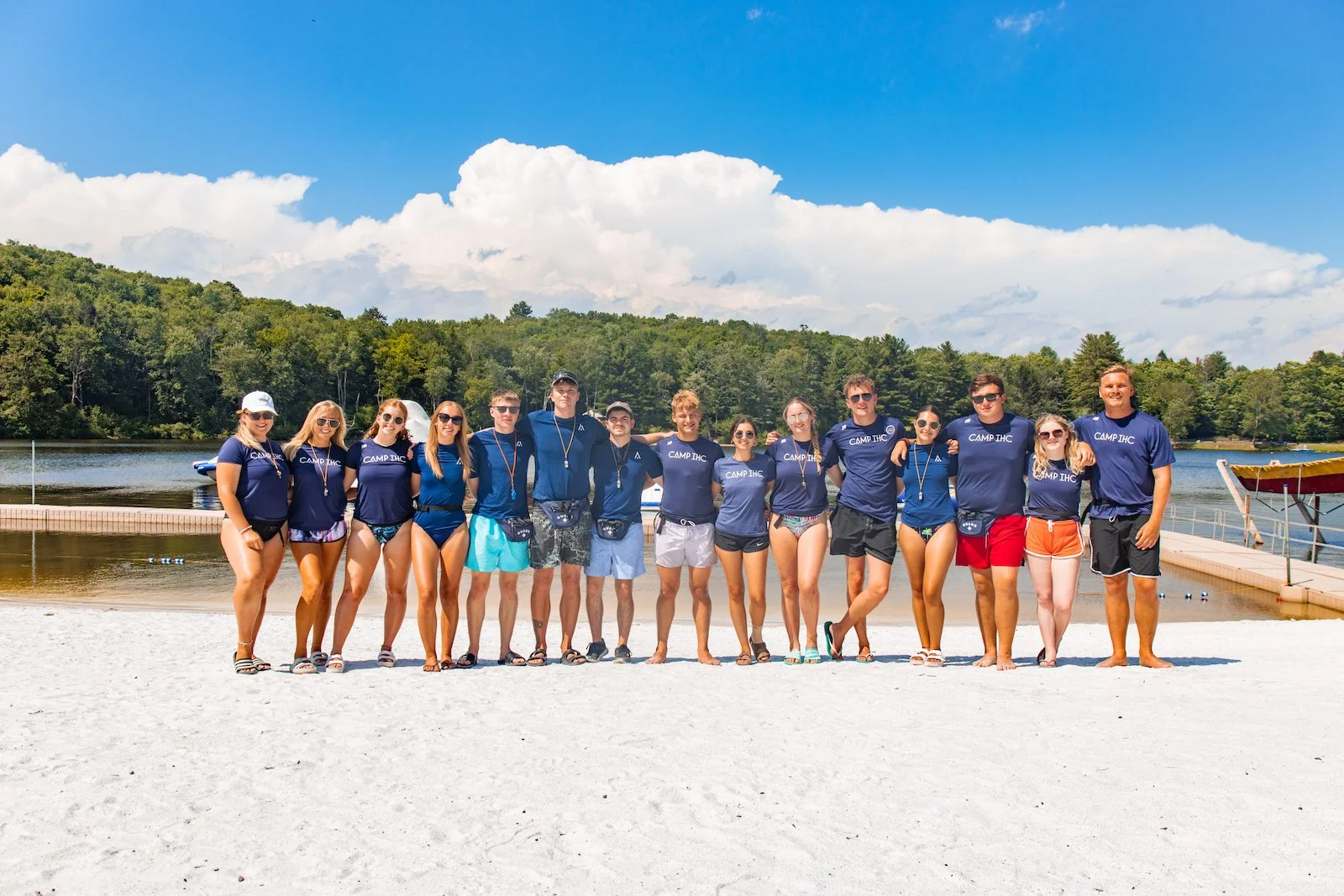 Group of teenagers in swimwear standing on a sandy beach with a lake, trees, and cloudy sky in the background.