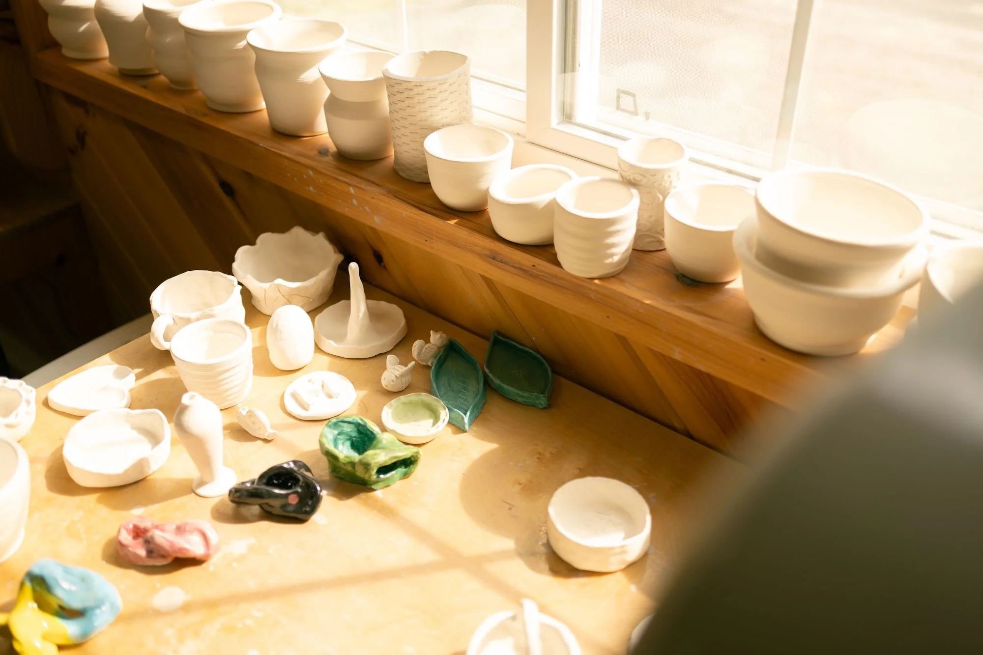 Assorted white pottery and ceramic pieces, including bowls, cups, and decorative objects, on a wooden table near a window.
