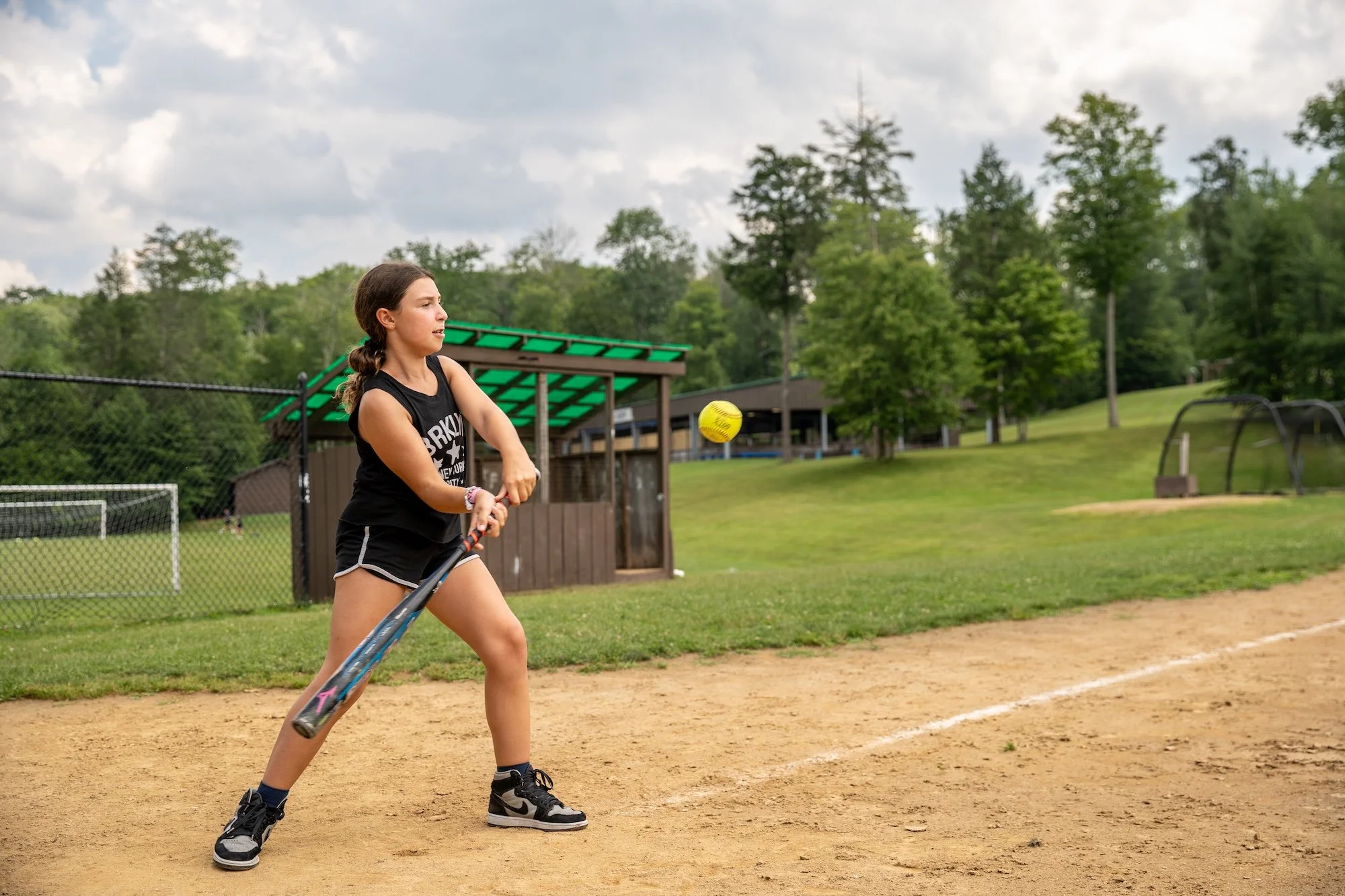 A girl playing softball on a dirt field, preparing to hit a yellow softball. She is wearing a black tank top, black shorts, and black and white sneakers. There are trees and a green-roofed shelter in the background.