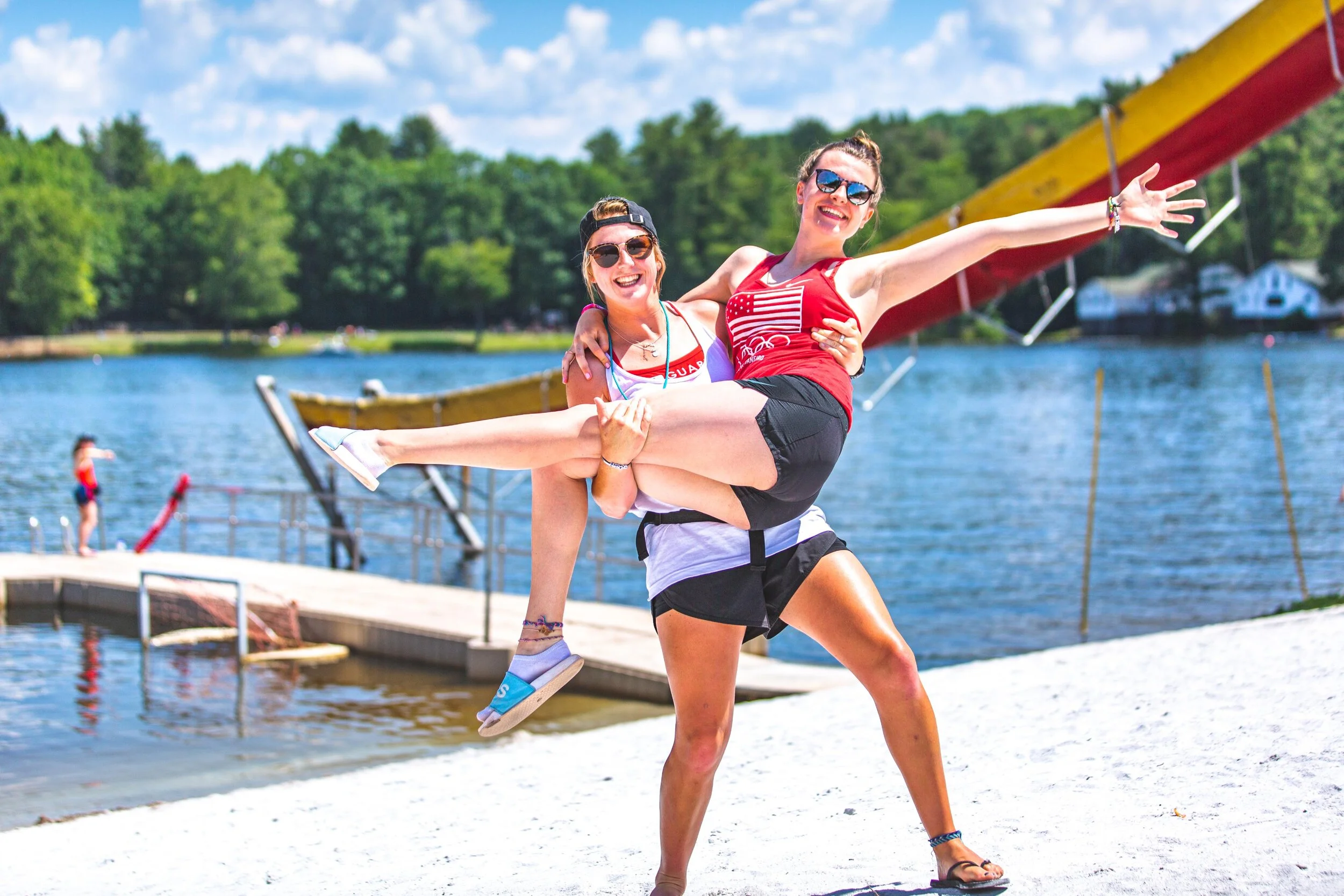 Two women smiling and posing by a lake, one holding the other in a playful pose, with a dock and a waterslide in the background on a sunny day.