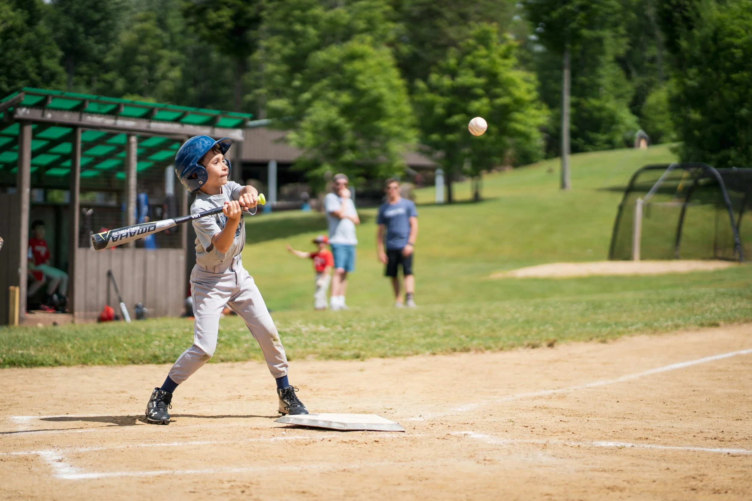 A young boy in a baseball helmet and uniform swings a bat at a pitched ball on a baseball field on a sunny day, with other children and adults in the background.