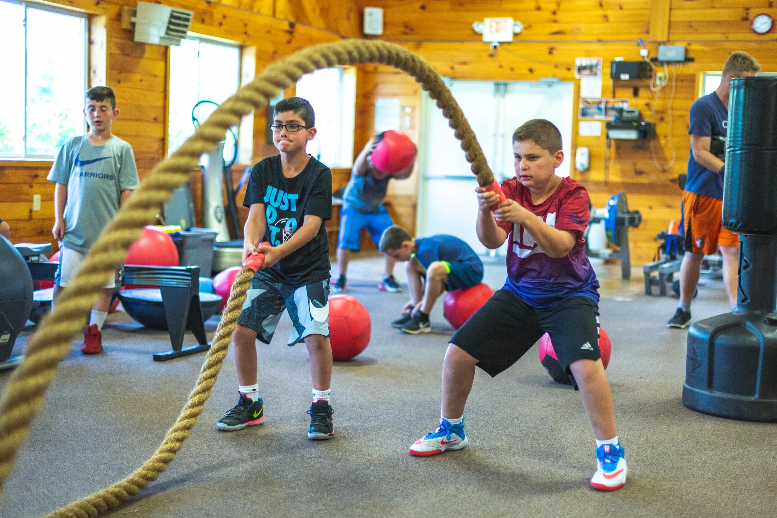 Group of boys working out in a gym, with some swinging battle ropes and others stretching or lifting exercise balls.