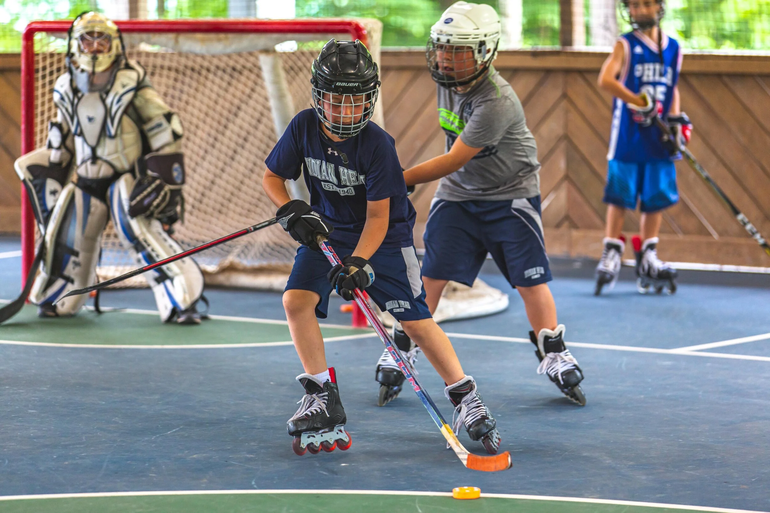 Two young boys playing street hockey on rollerblades indoors, with one controlling the puck and the other defending, while a goalkeeper in full gear stands behind them, and another child watches in the background.