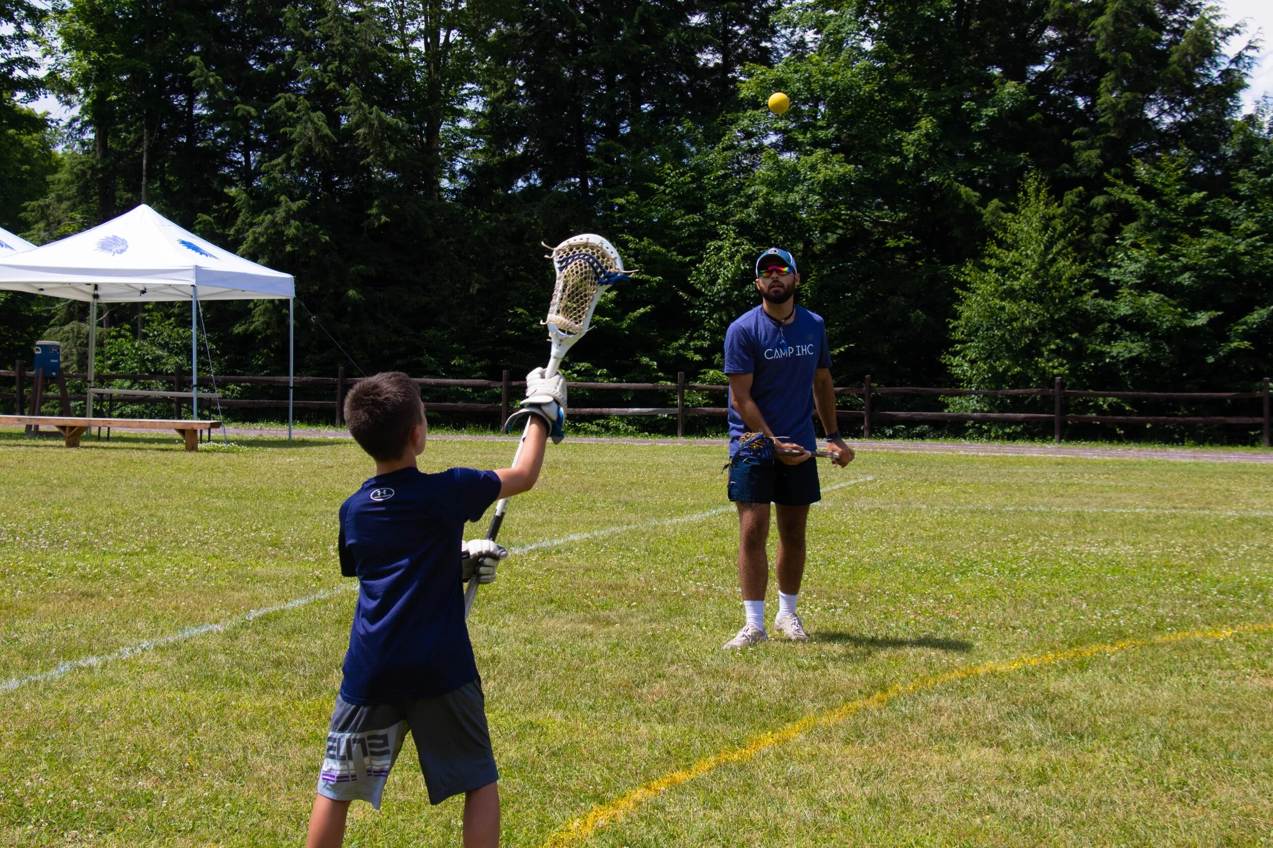 A boy playing lacrosse with a man watching on a grassy field surrounded by trees and tents.