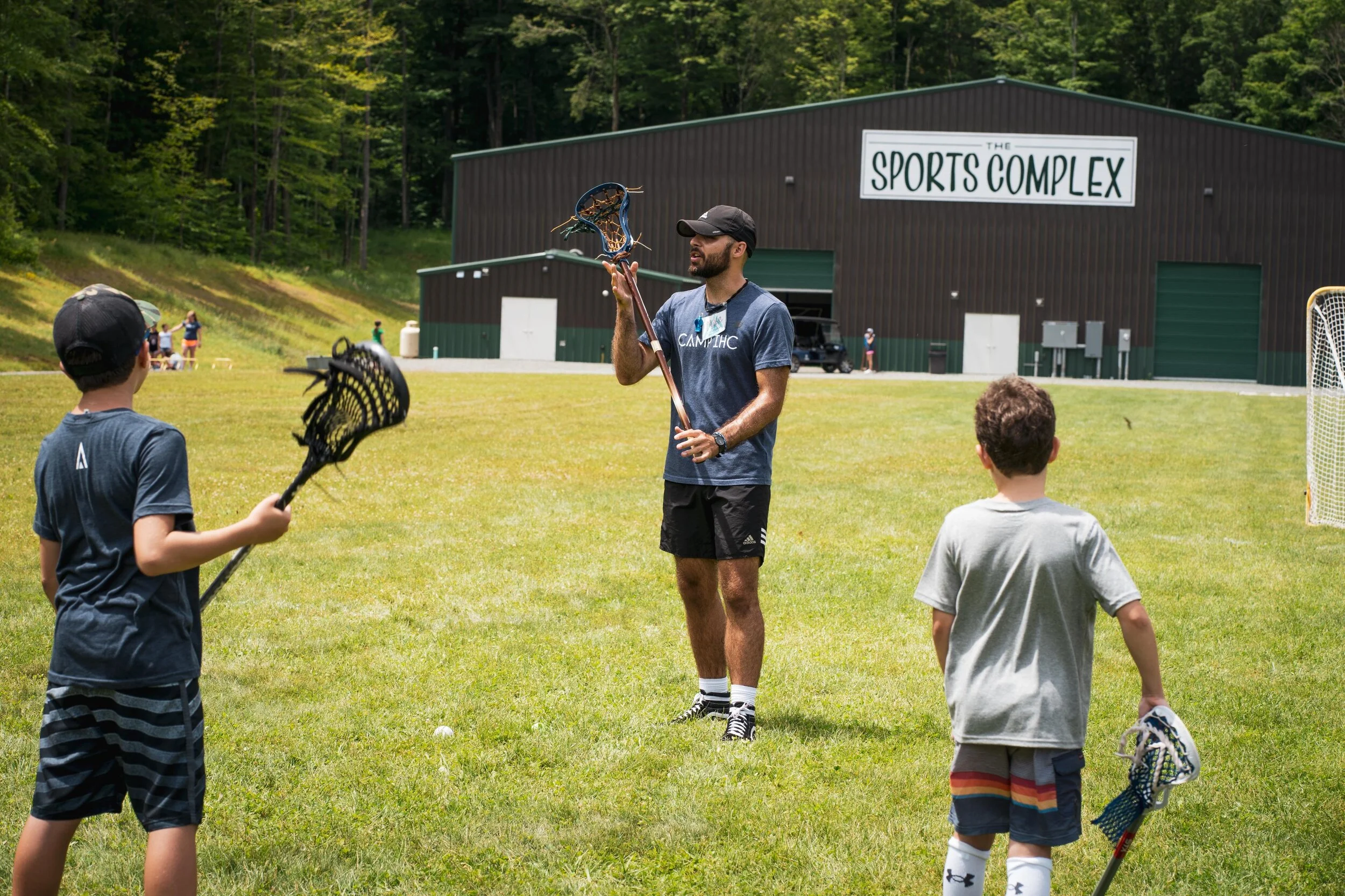 A man in athletic clothing demonstrating lacrosse to two young boys on a grassy field outside a sports complex building with a sign that reads 'The Sports Complex'.