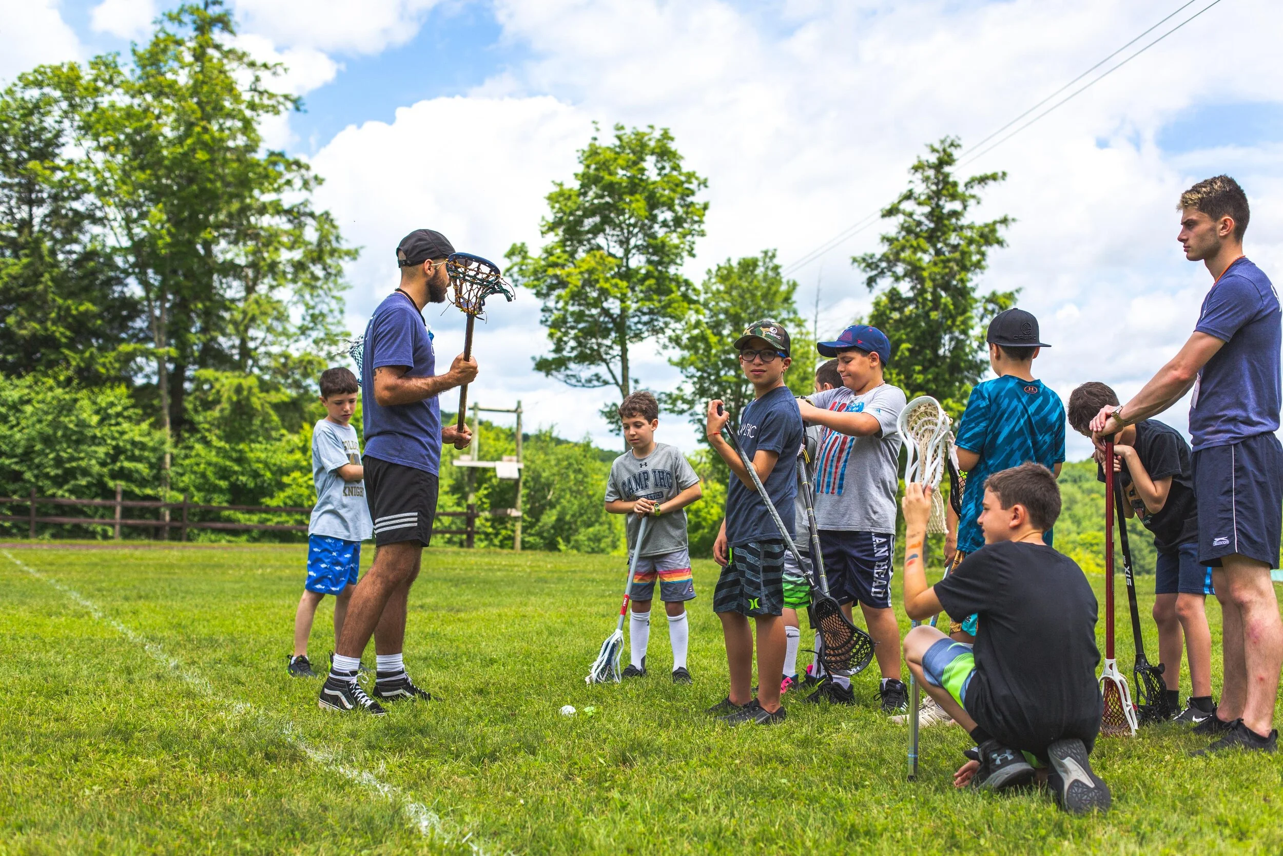 A group of young boys and two coaches gathered on a grassy field for a lacrosse practice or lesson, with one coach holding a lacrosse stick and explaining something to the kids, some holding lacrosse sticks, outdoors on a sunny day with trees in the