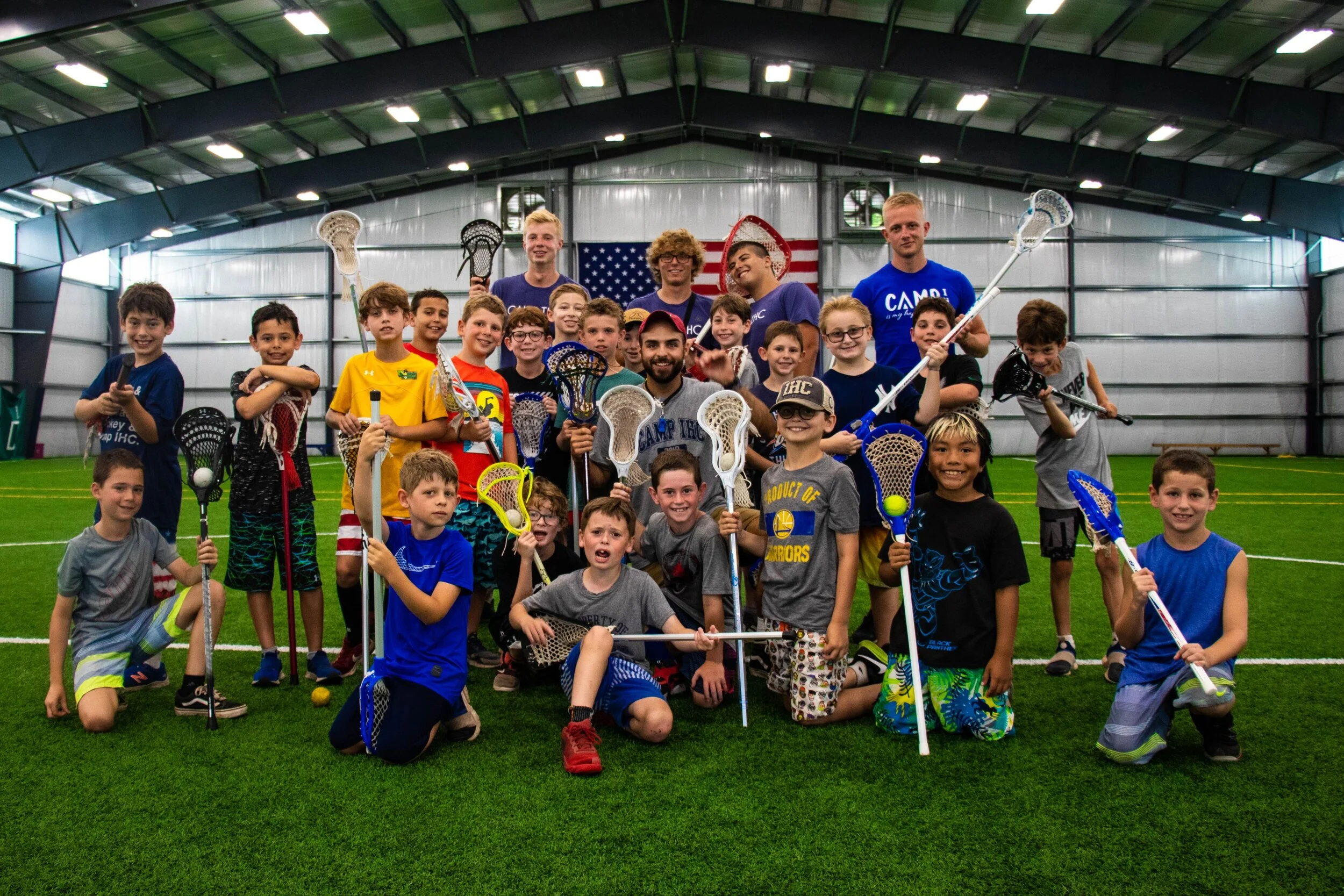 Group of children and adult coaches in an indoor lacrosse training facility, all holding lacrosse sticks and smiling, with an American flag in the background.