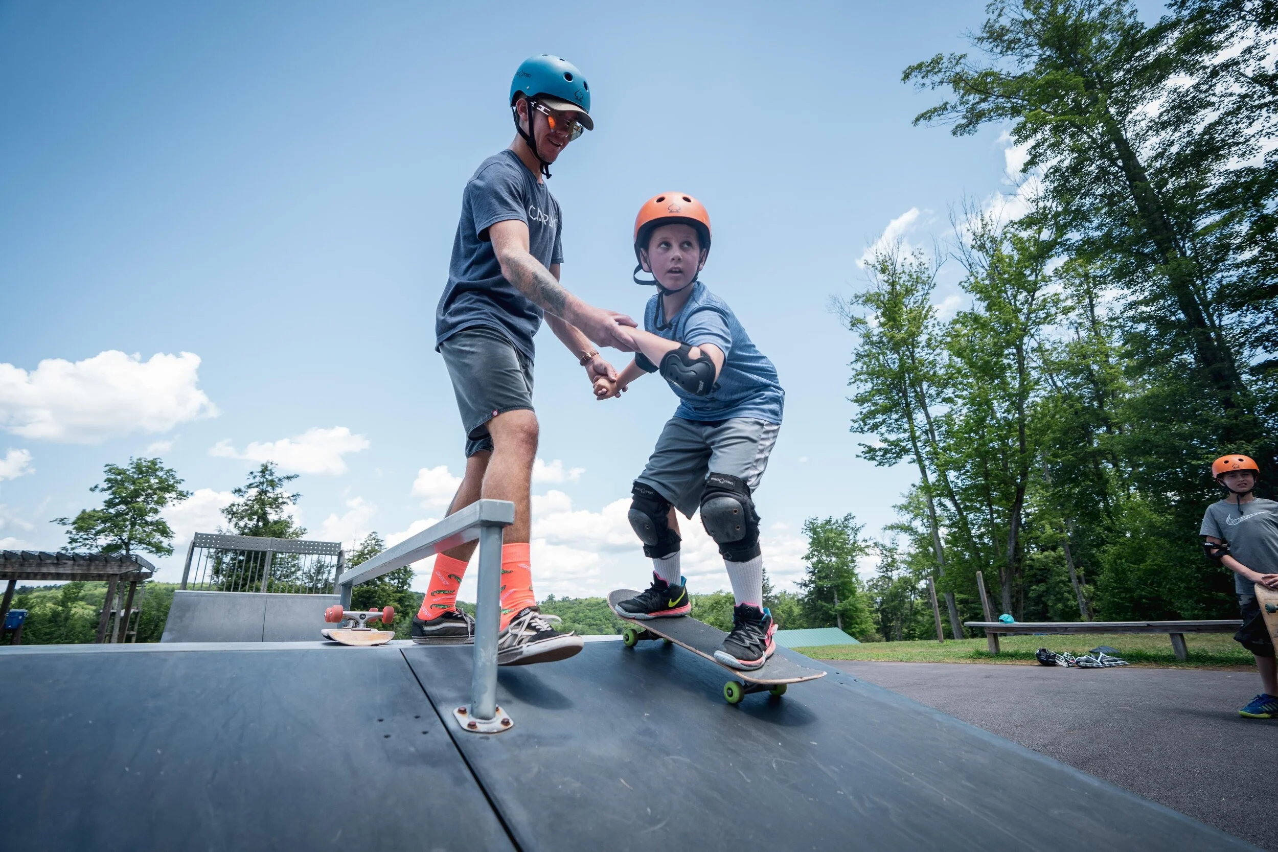 A man helping a young boy skateboard at a skate park during the daytime, both wearing helmets and knee pads, with trees and a clear sky in the background.