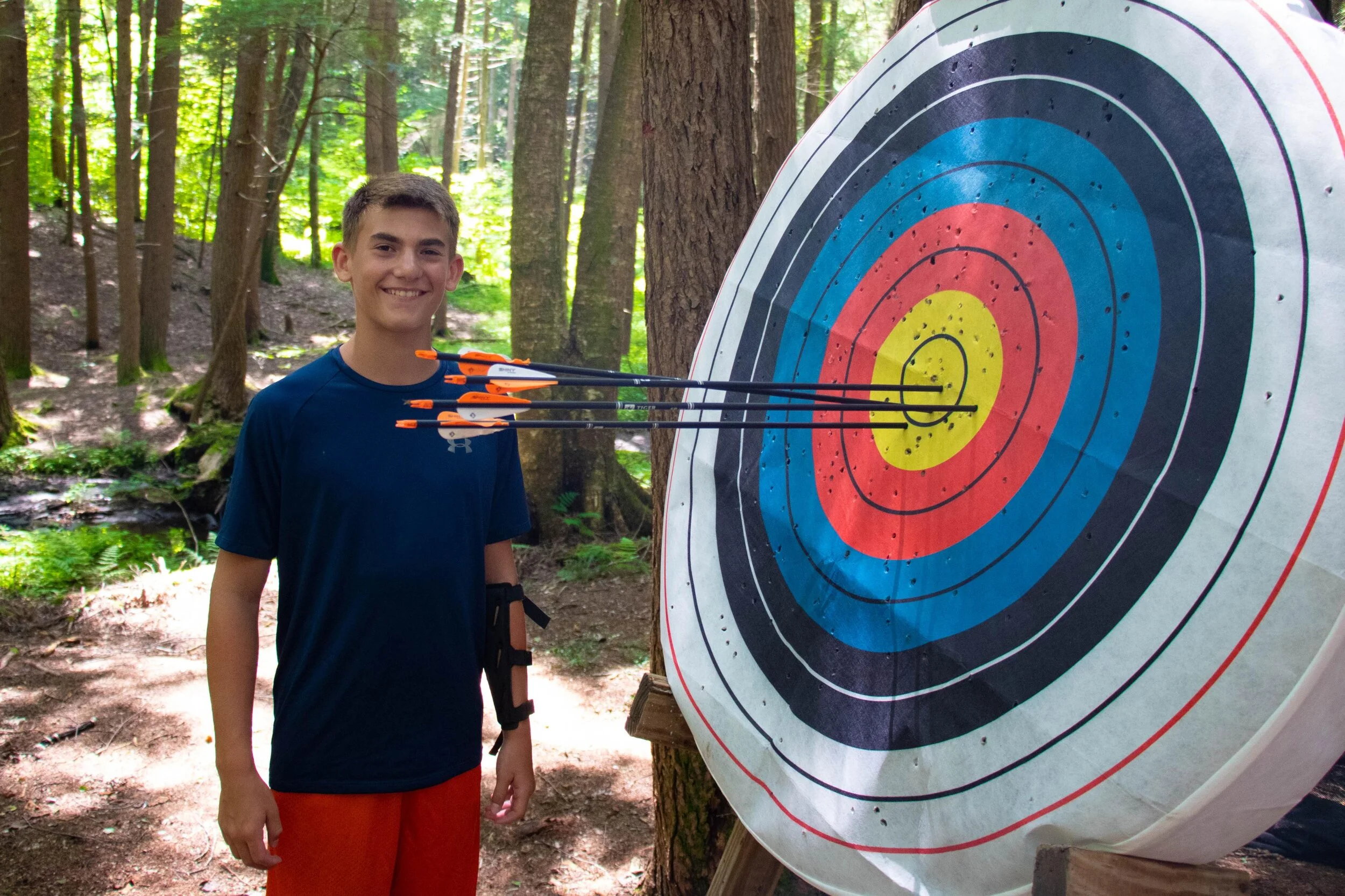 A young boy with short brown hair, wearing a navy blue t-shirt and red shorts, stands in a forested area smiling at the camera with four arrows embedded in the bullseye of a large archery target.
