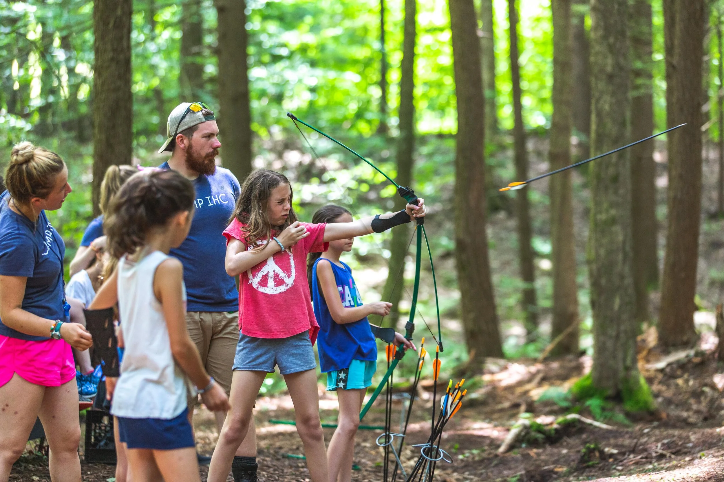 A group of children and an adult in a forest participating in archery, with arrows on a stand and one girl aiming and shooting an arrow.