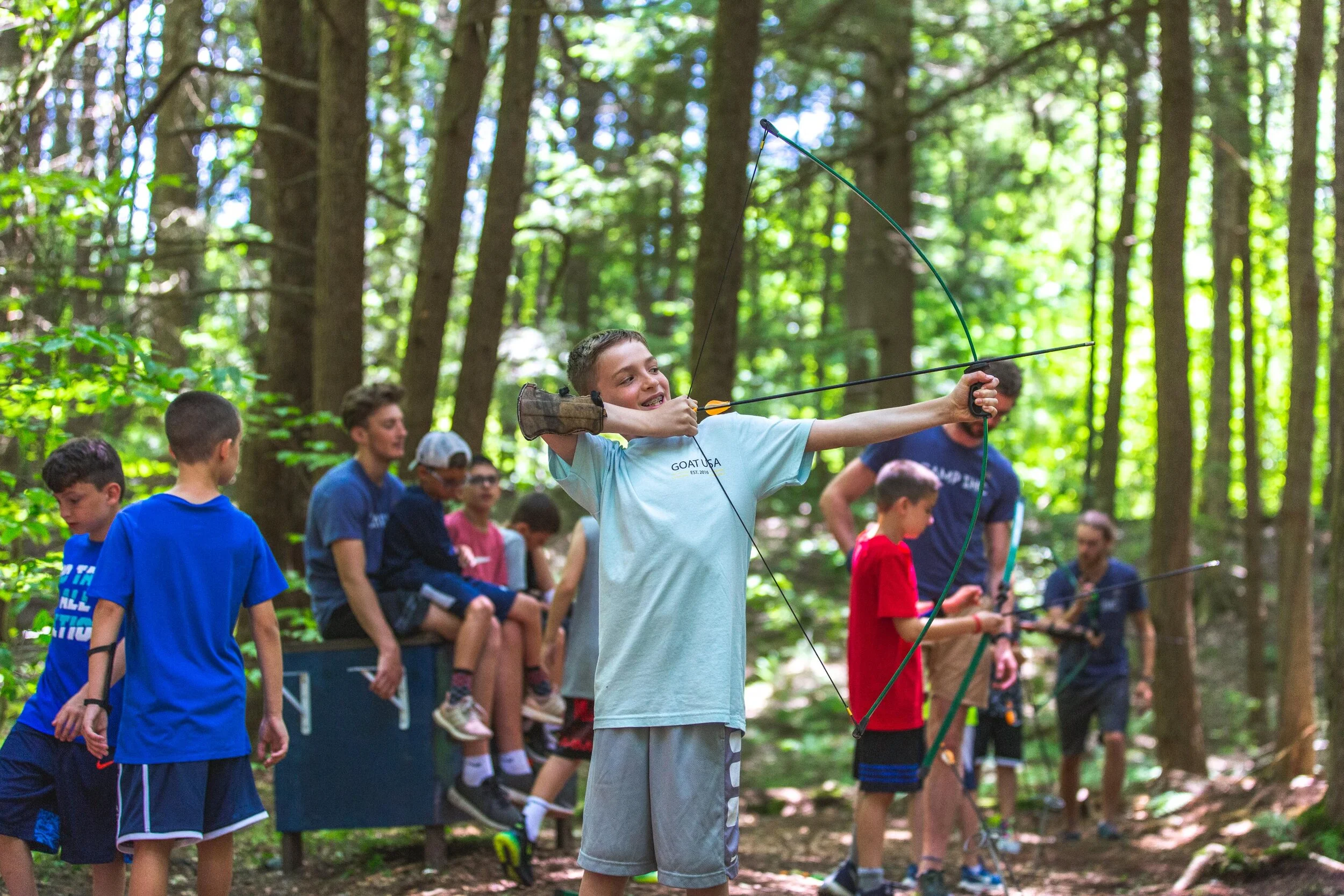 Boy aiming a bow and arrow in a forest, with other children and adults watching nearby.