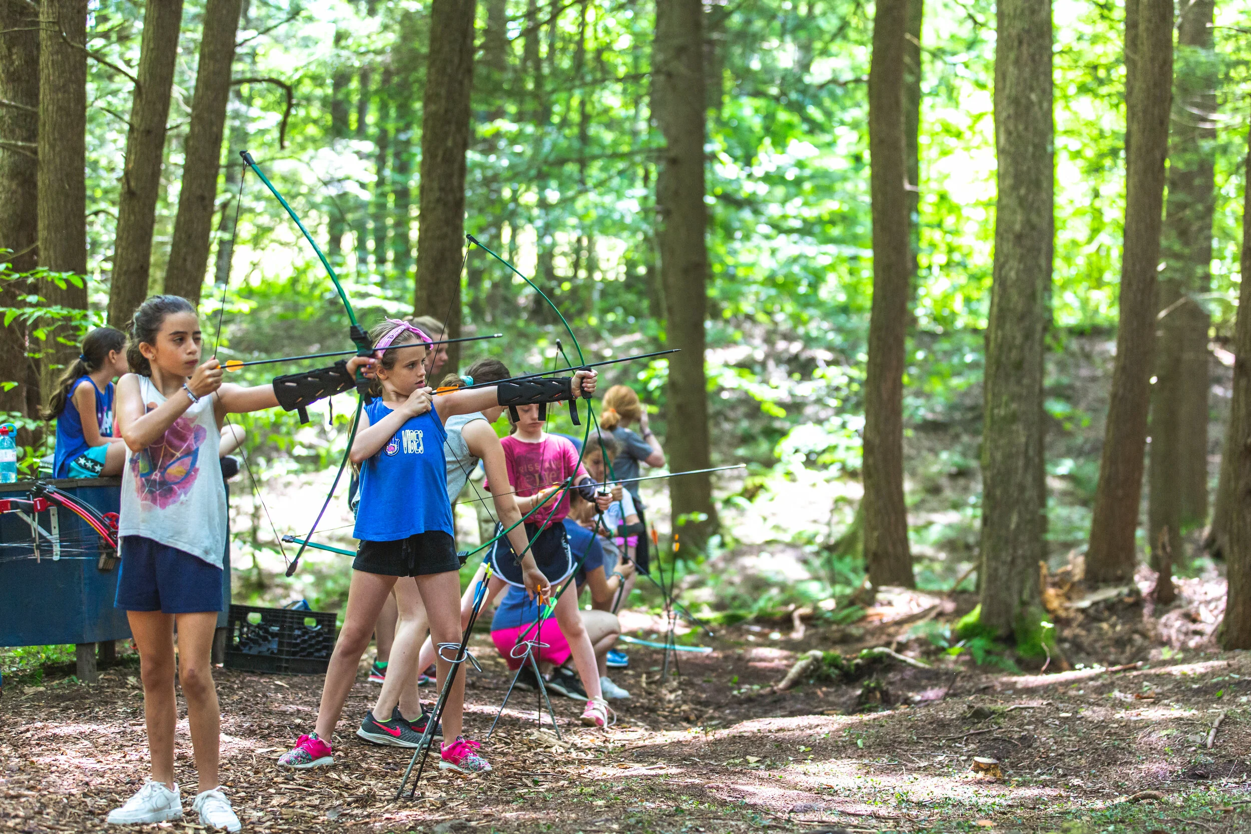 A group of young girls practicing archery in a wooded forest, some aiming bows and arrows while others observe.