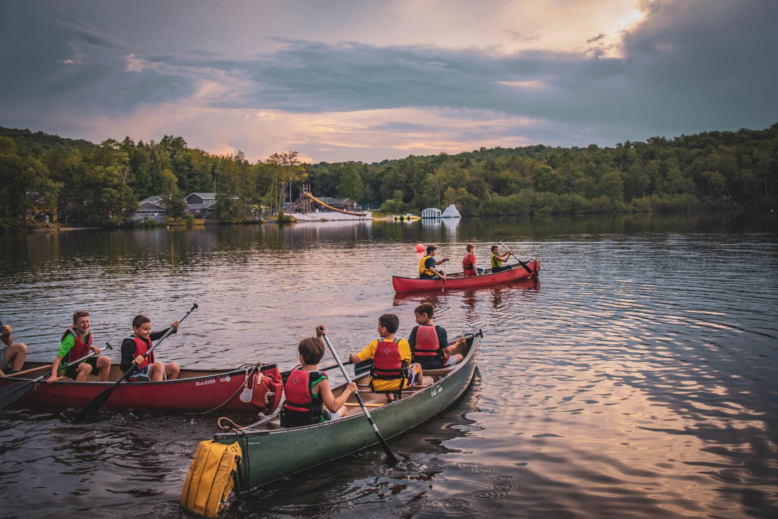 Children in colorful canoes paddling on a calm lake at sunset, with trees and lakeside houses in the background.