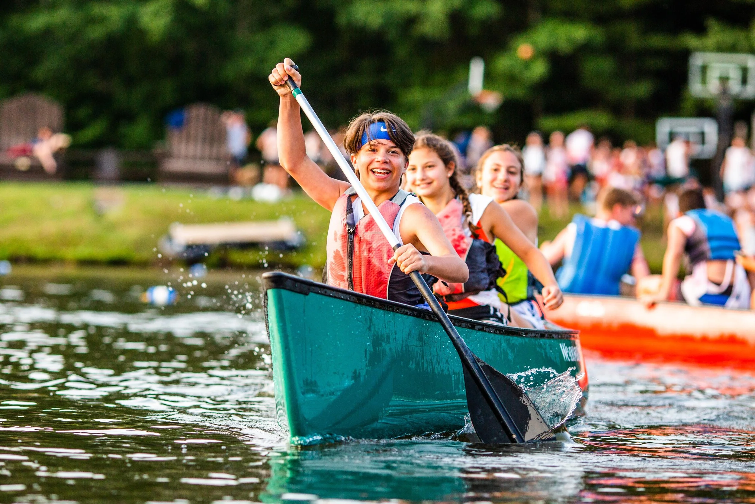 Children enjoying a boat ride on a lake with two girls paddling a canoe in the foreground and other children in the background, all wearing life jackets and smiling.