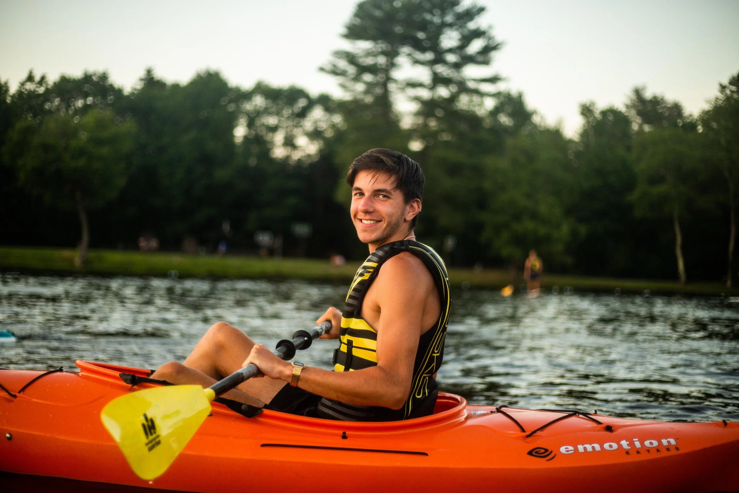 A smiling young man kayaking on a lake during the day, wearing a life jacket and holding a paddle.