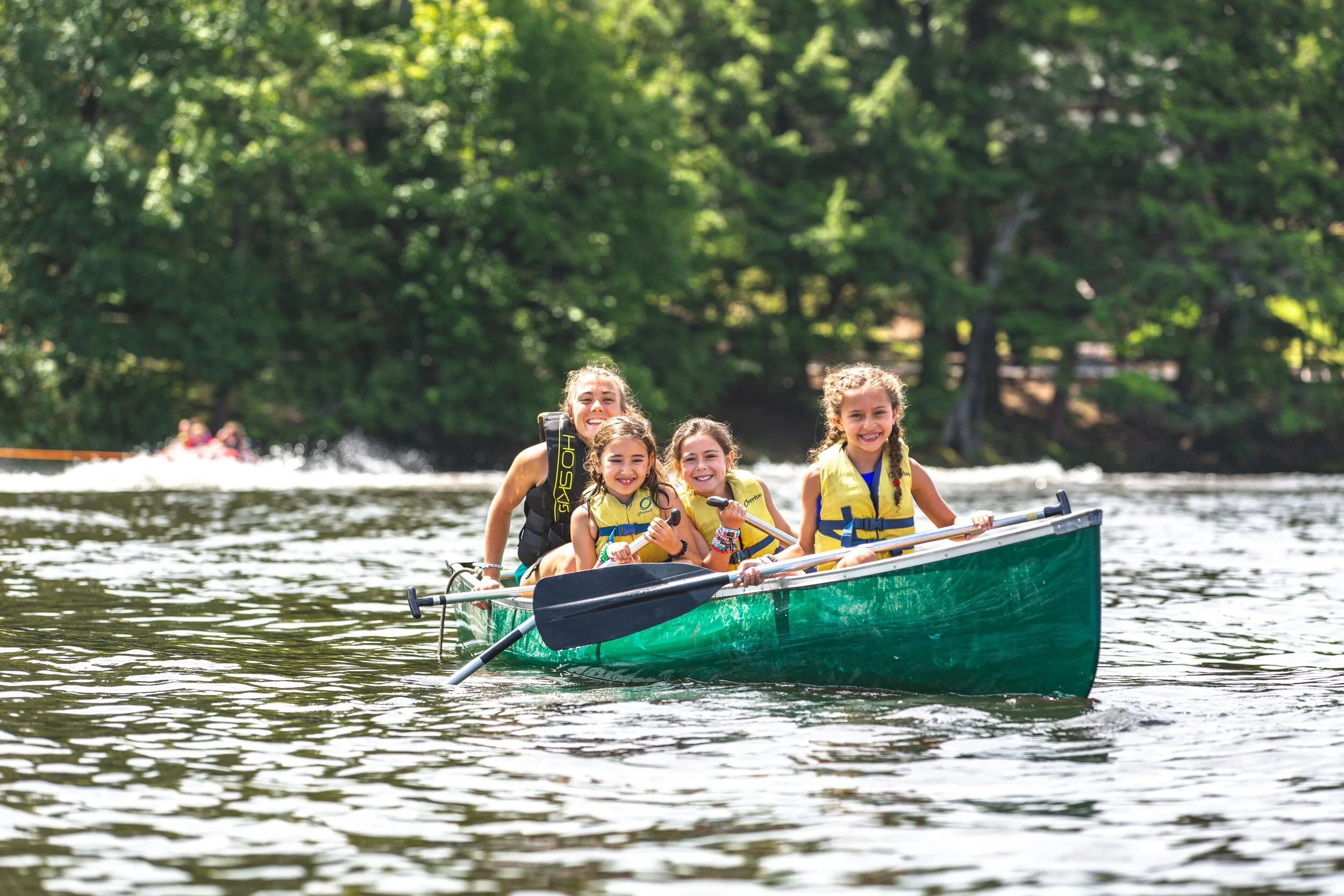 Four children wearing life jackets and paddling a green canoe on a lake surrounded by green trees, smiling and enjoying a sunny day.